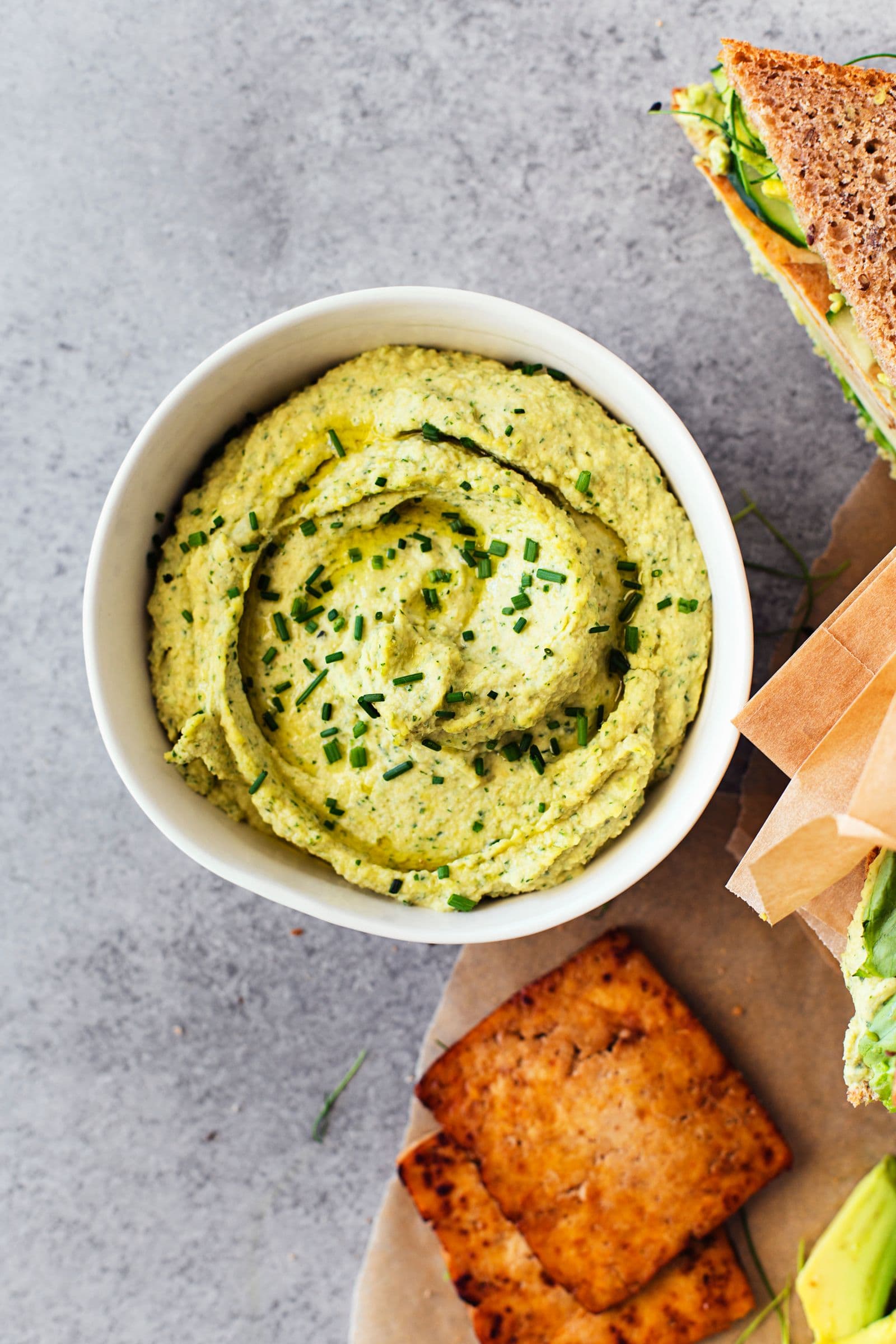 Creamy green herb hummus in a small white bowl with sandwich and tofu on the side.
