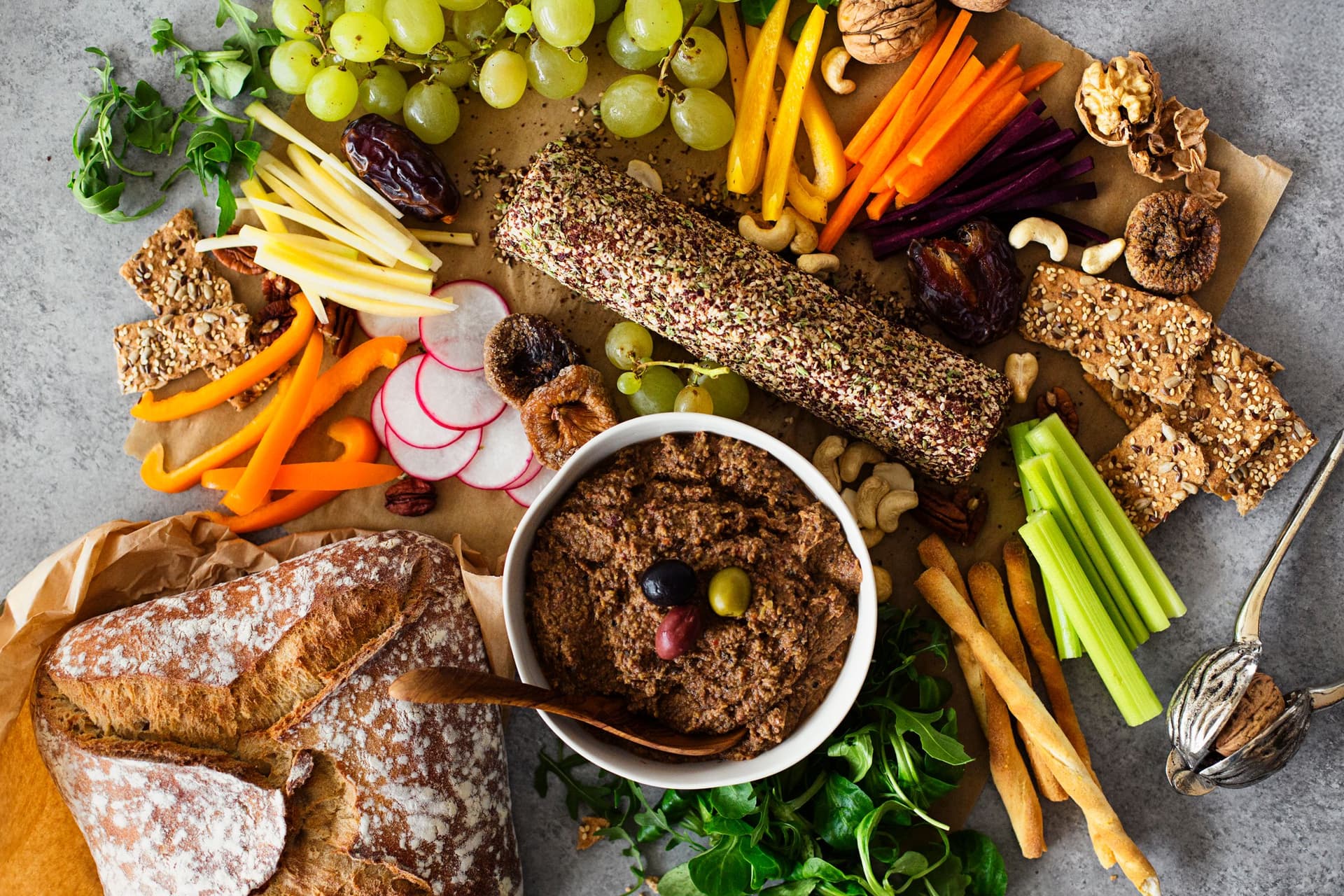 Festive platter with a cashew cheese log coated in herbs, a bowl of lentil and olive pâté, fresh grapes, vegetables and slices of bread.