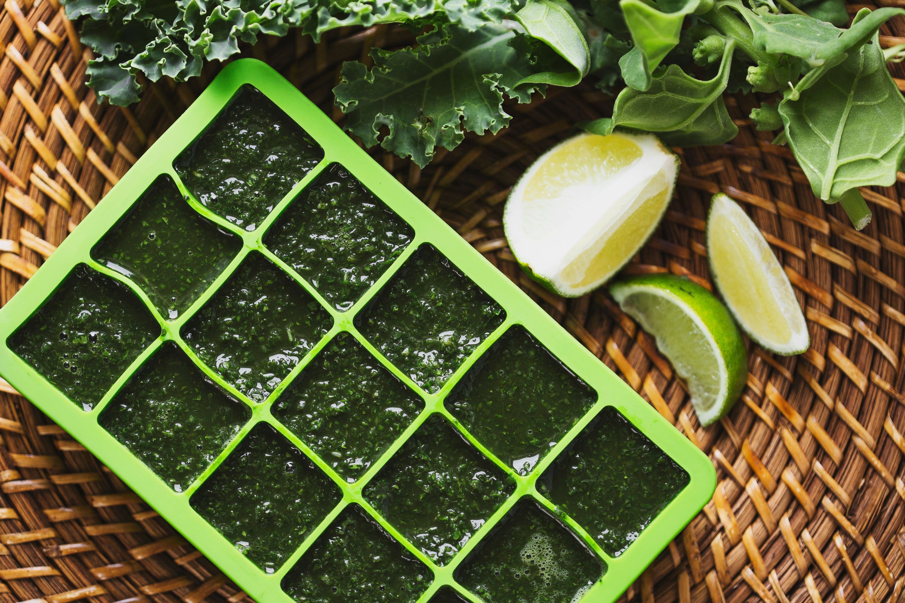 Tray of green smoothie cubes surrounded by fresh greens and lime on a rattan tray.