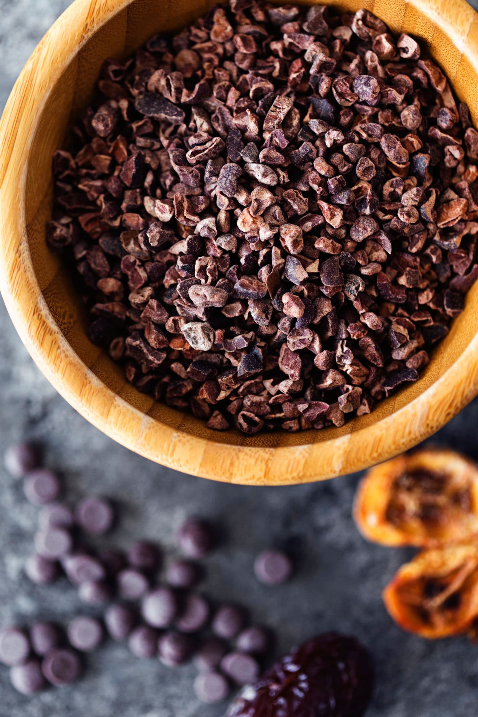 Wooden bowl with cacao nibs next to banana chips and dark chocolate chips.