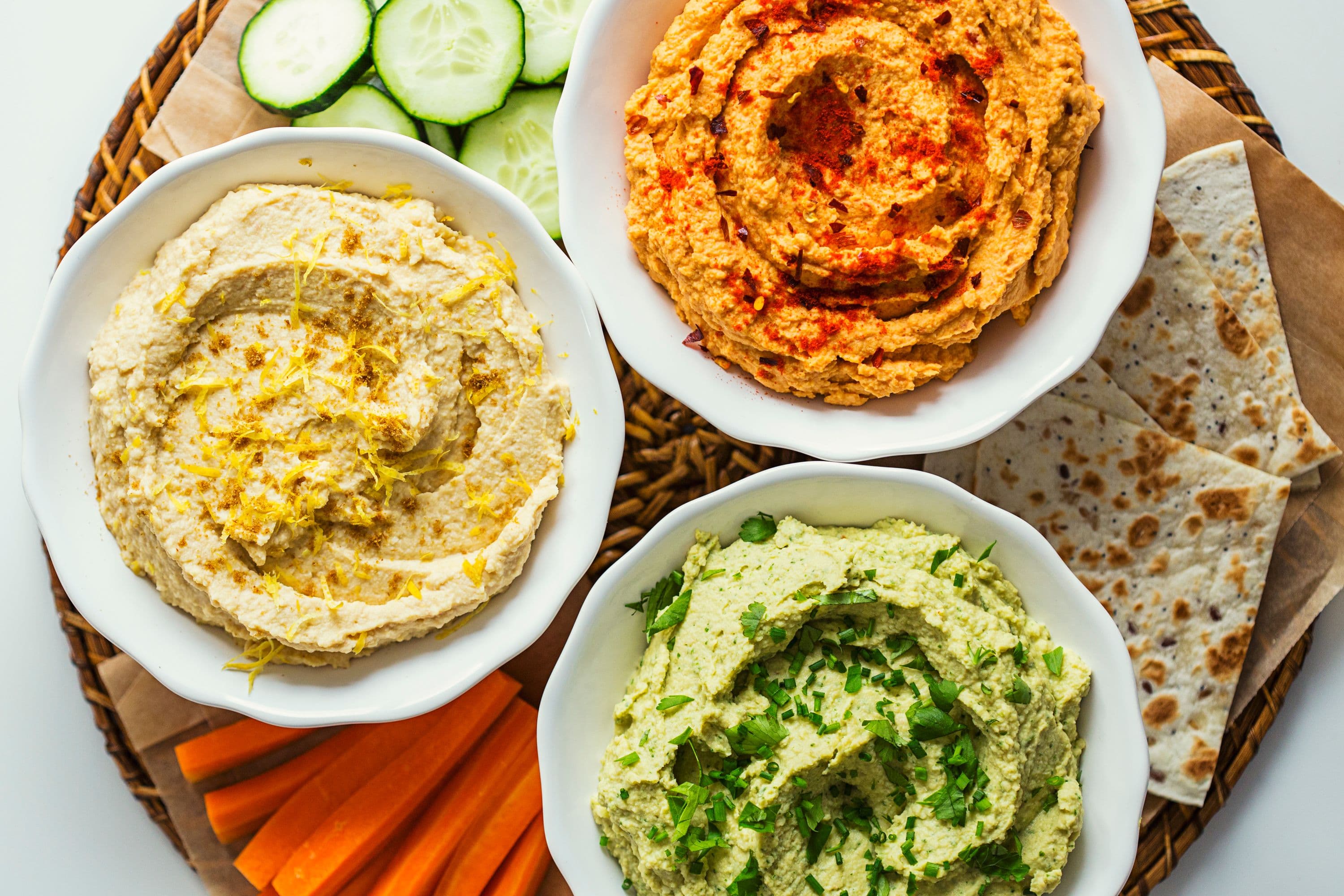 Three colourful bowls of hummus—lemon, tomato, and herb—served with fresh tortilla and vegetable sticks.