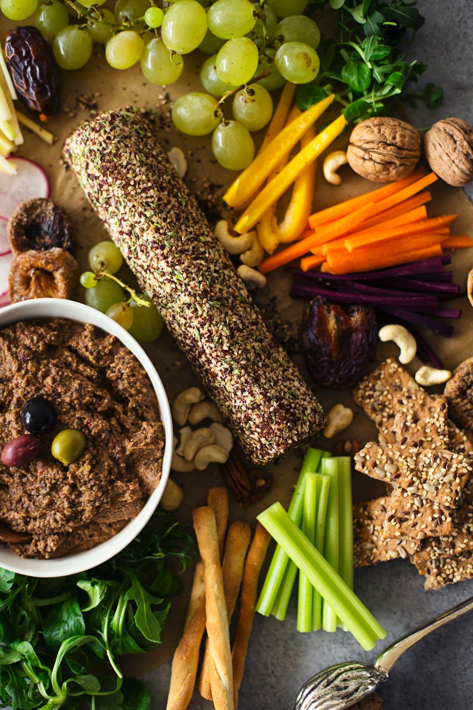 Cashew cheese log and lentil pâté served on a large platter with grapes, nuts, raw vegetables and bread slices.