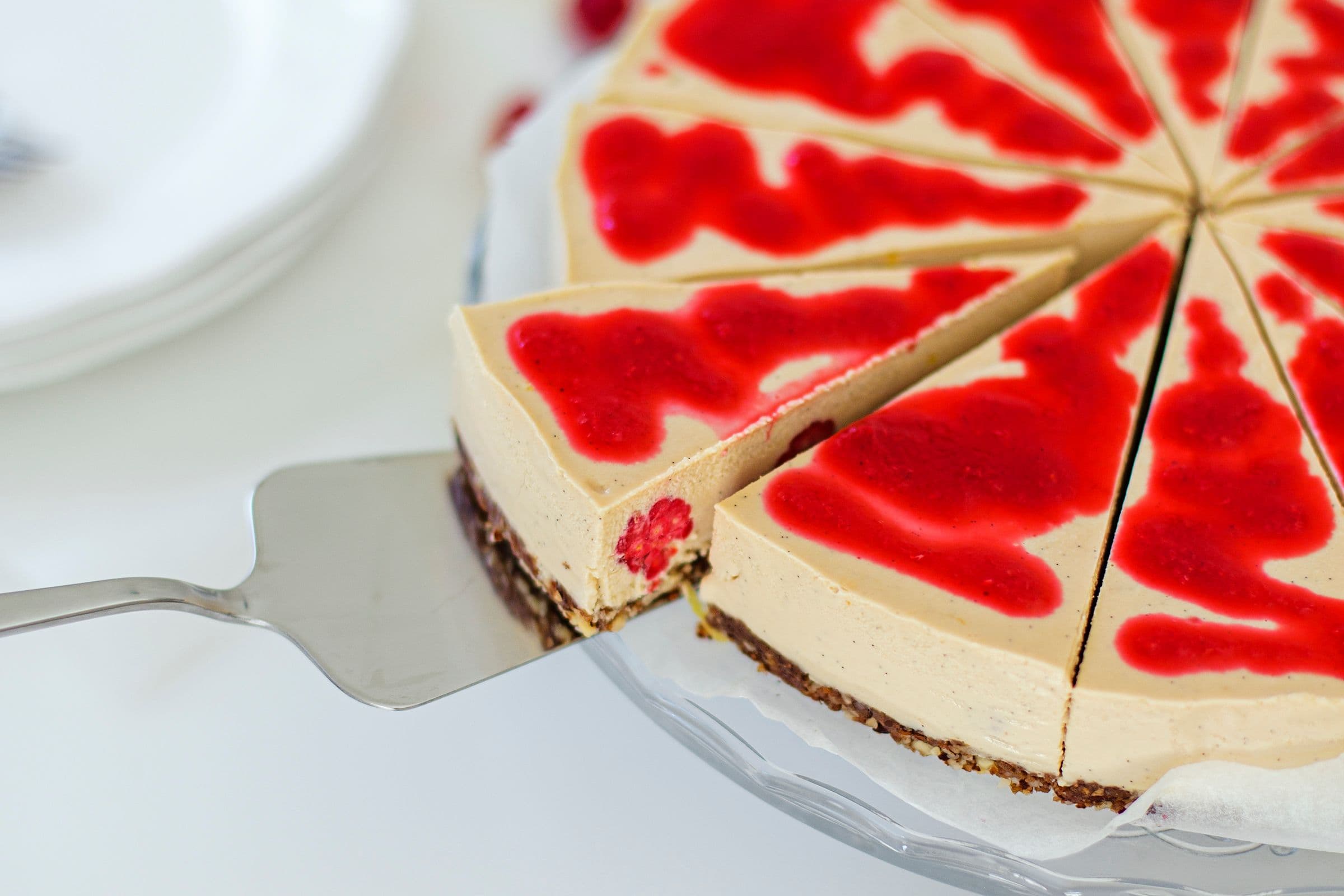 Cheesecake vegan fatiado com molho de framboesa, servido num prato de bolo.