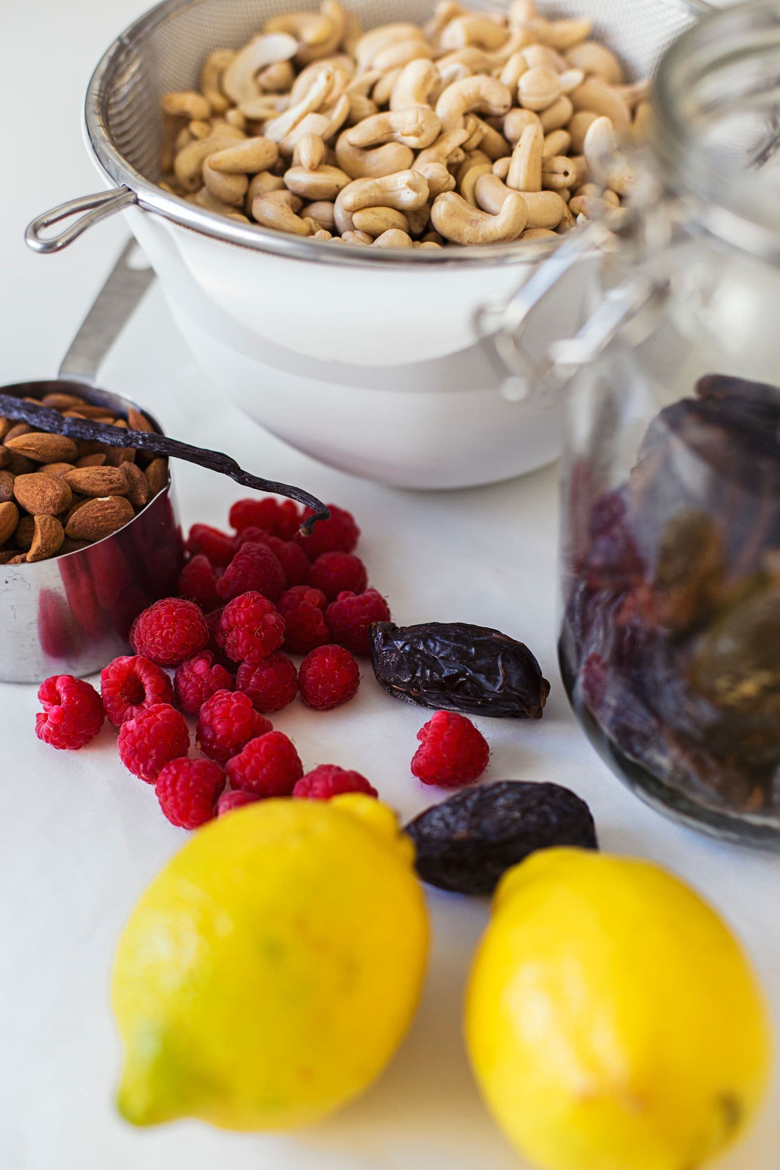 Cashews, raspberries, lemons, dates and vanilla beans on a white surface.