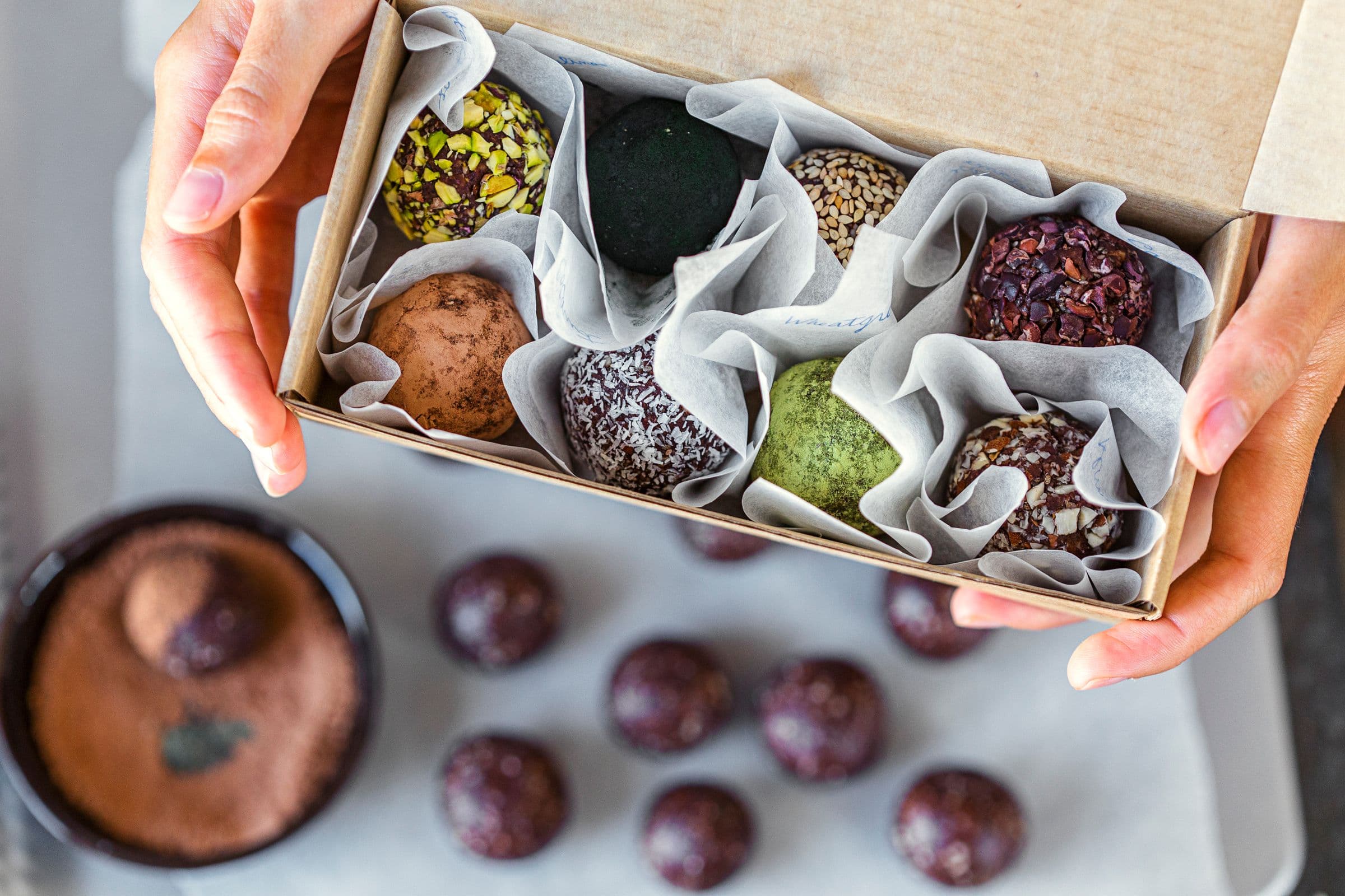 Hands holding a neatly packed gift box with energy balls rolled in colourful toppings.
