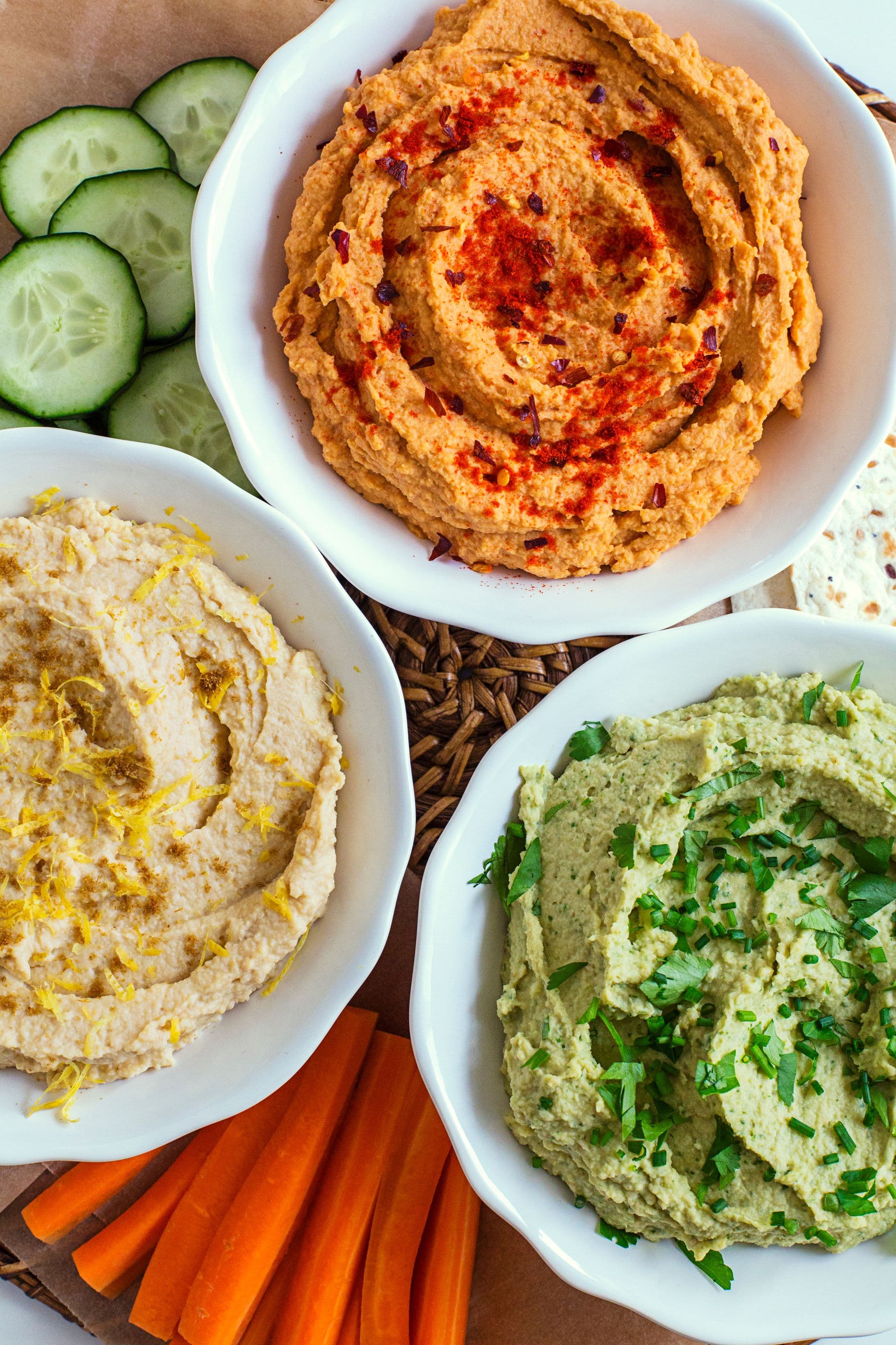 Three finished hummus bowls with garnishes, served with cucumber slices and carrot sticks.
