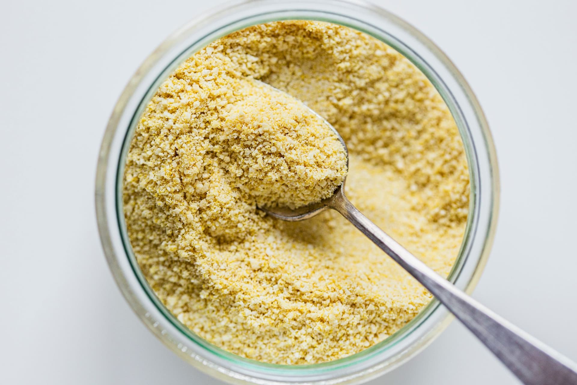 Spoon scooping up finely ground vegan parmesan cheese from a jar.
