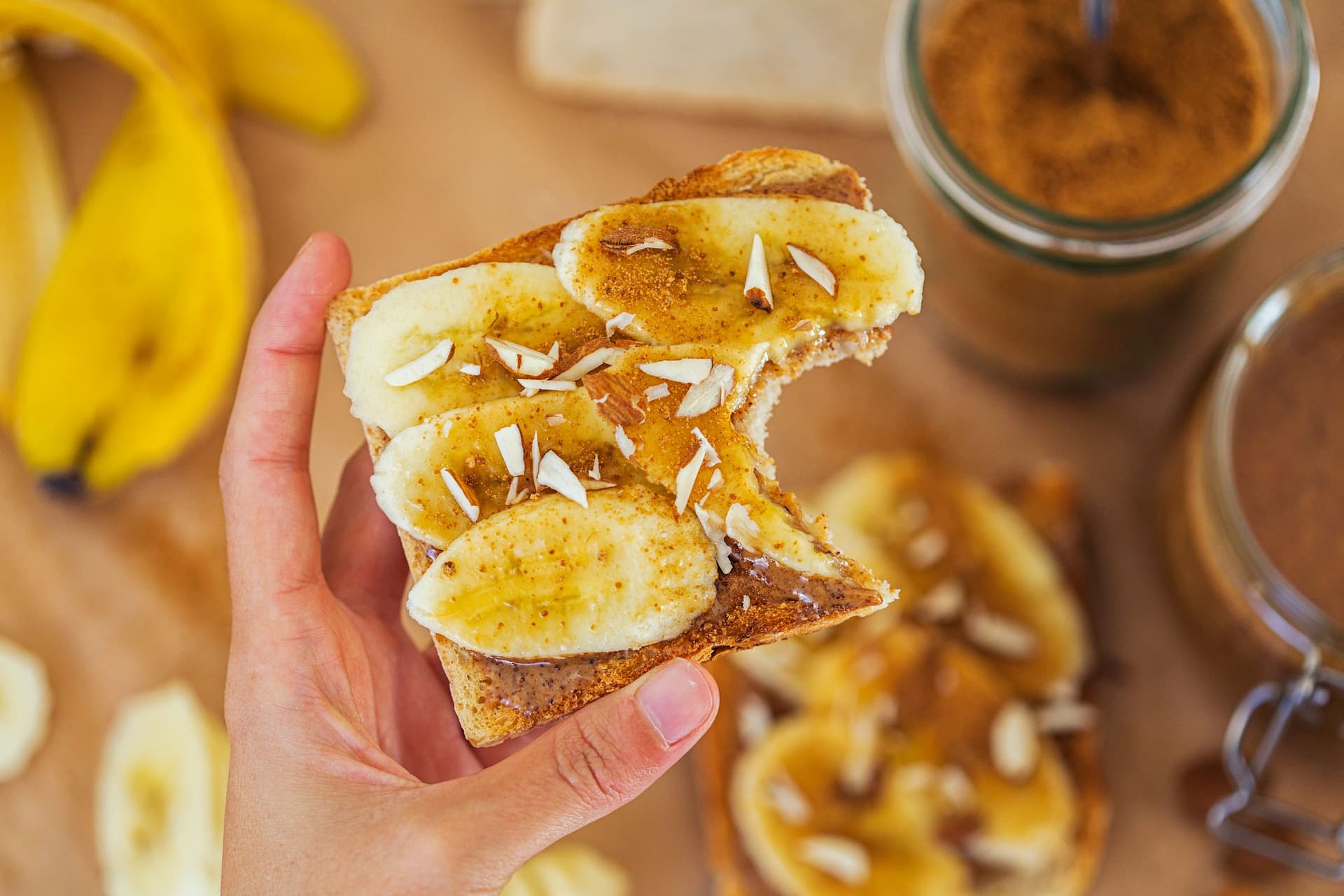 Mão a segurar uma fatia de pão com manteiga de amêndoa, banana e amêndoas picadas.