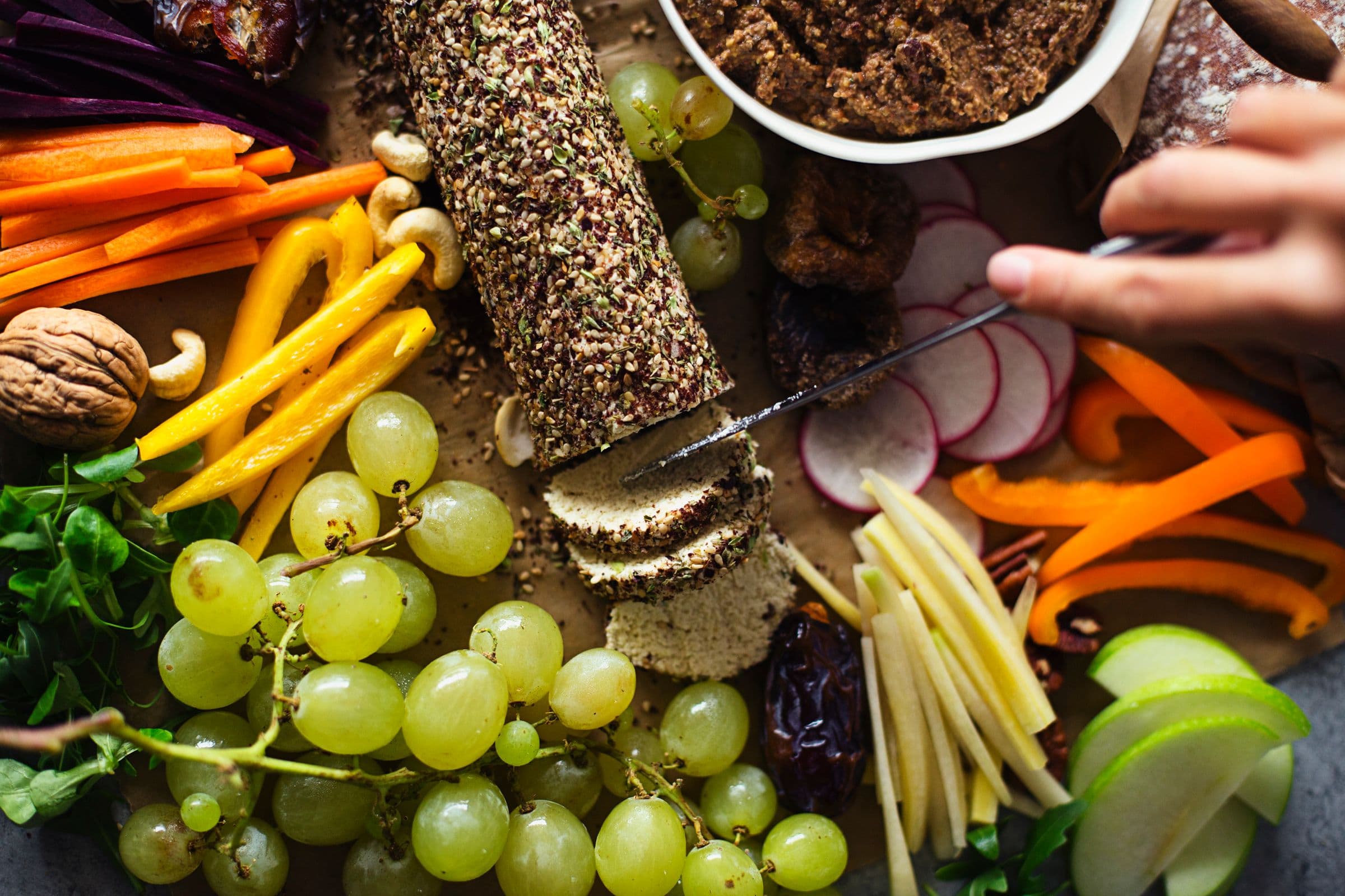 Hand slicing cashew cheese log on a platter surrounded by green grapes and fresh vegetables.