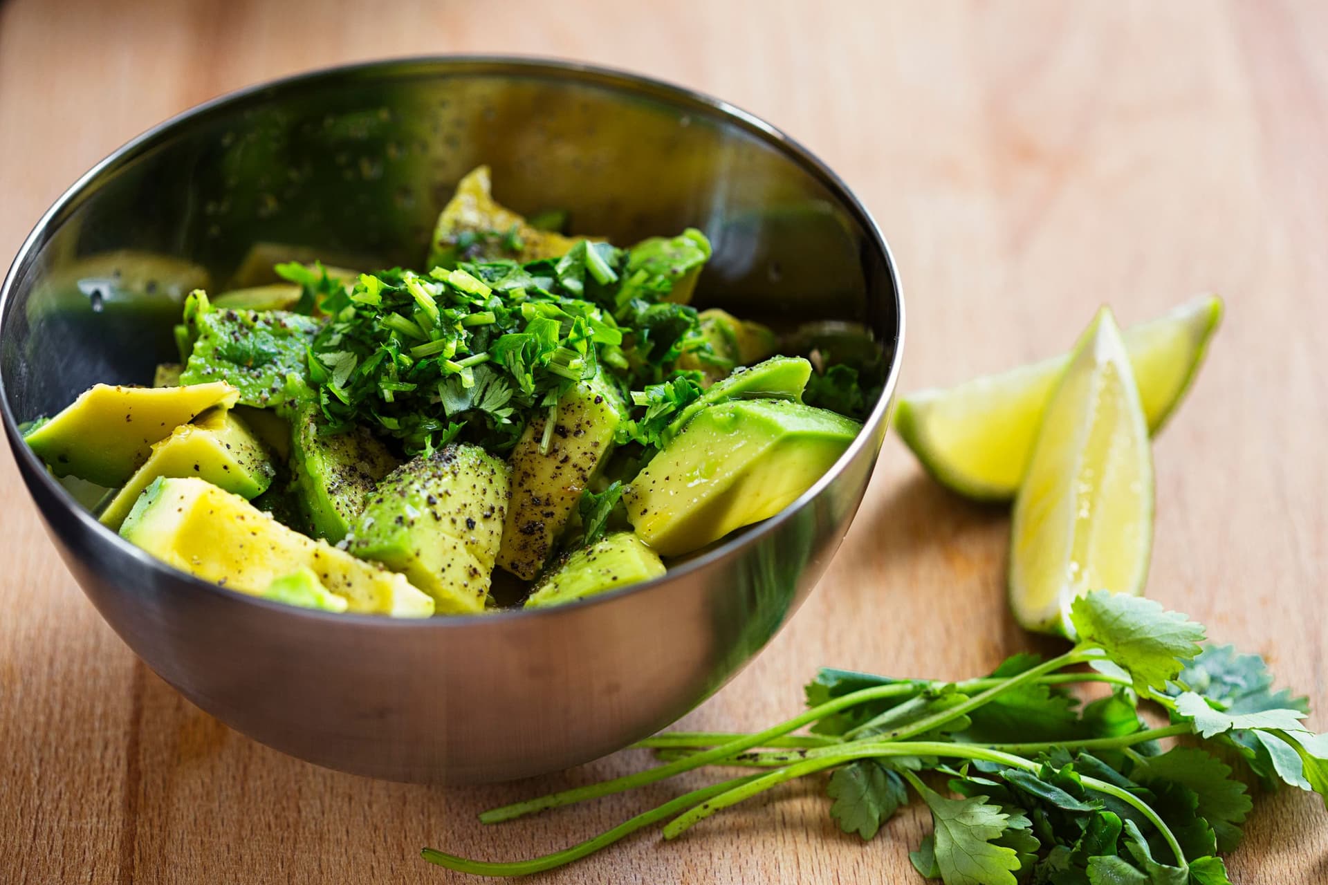 Metal bowl with avocado chunks, coriander and spices on a wooden surface with lime wedges.
