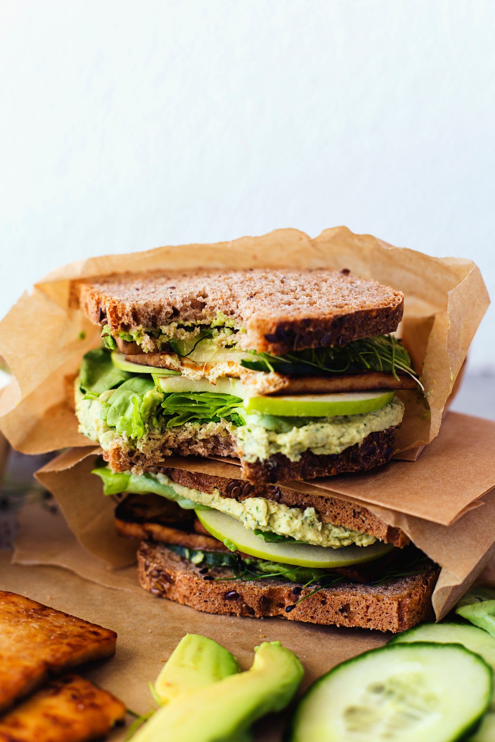 Stacked green sandwich with tofu, lettuce, cucumber and hummus on parchment paper, with a bite taken off.