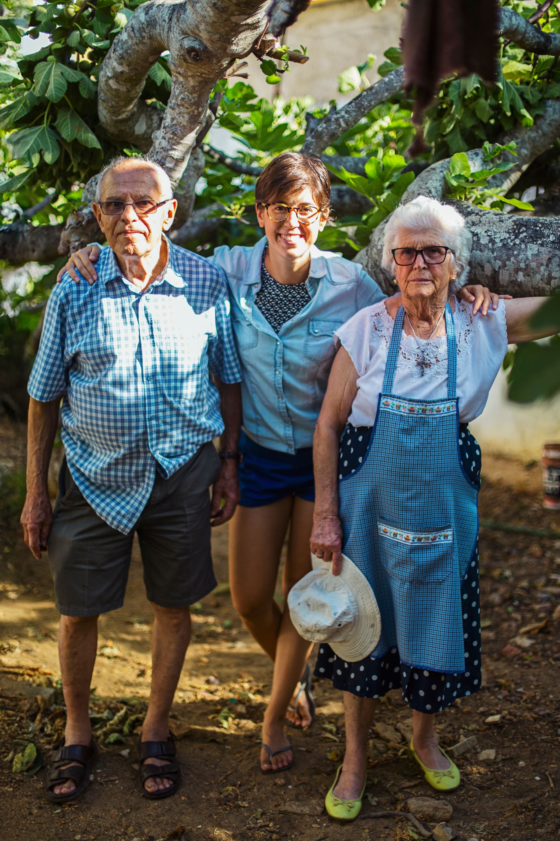 Portrait of Rita with her grandparents, standing in the fig orchard smiling.