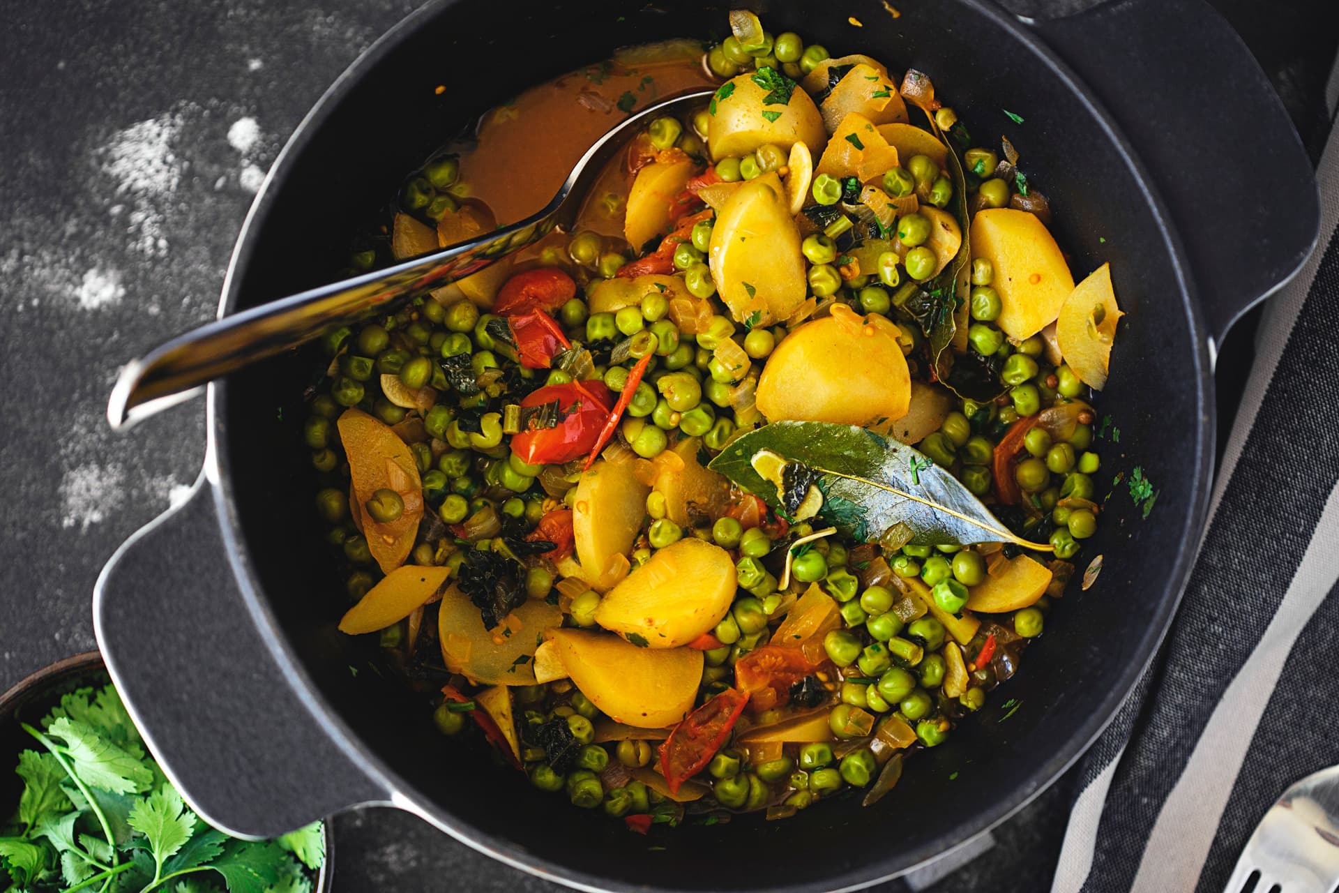 Close-up of vibrant vegan pea stew in a black pot with potatoes, cherry tomatoes and bay leaf.