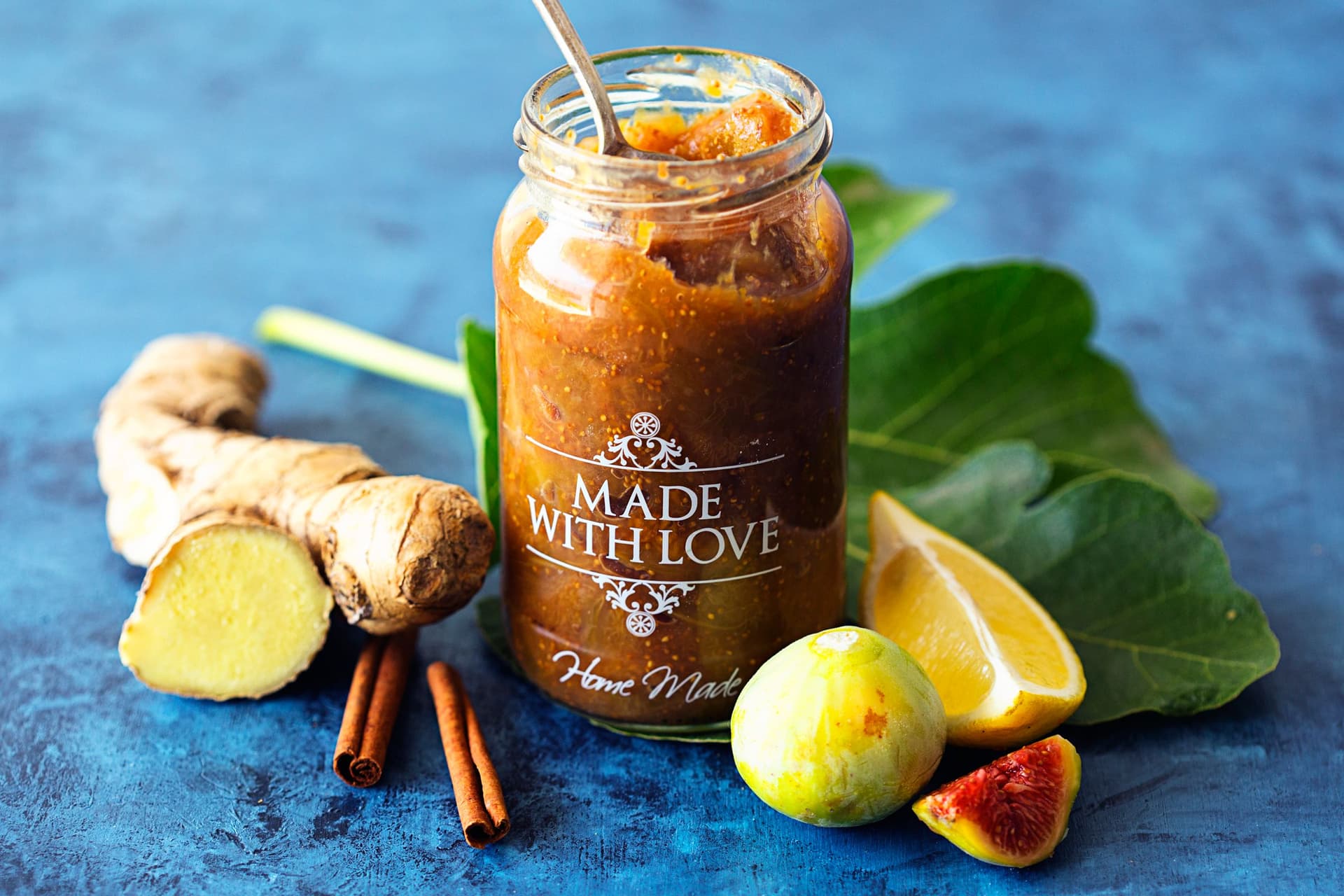 Jar of fig and ginger jam with ginger root, lemon, cinnamon sticks and fig leaf in the background.