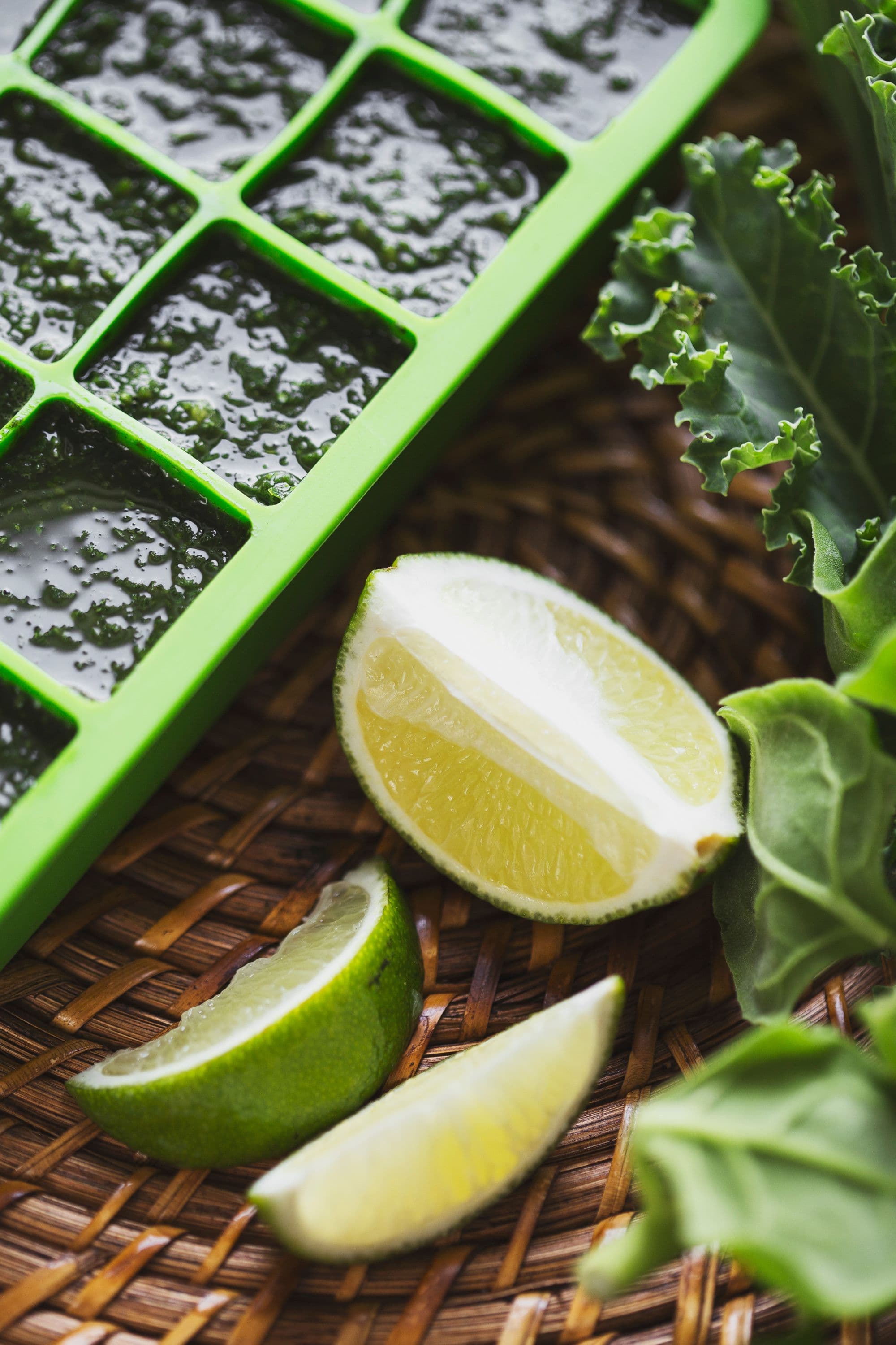 Green ice cube tray filled with blended greens next to lime wedges and kale.