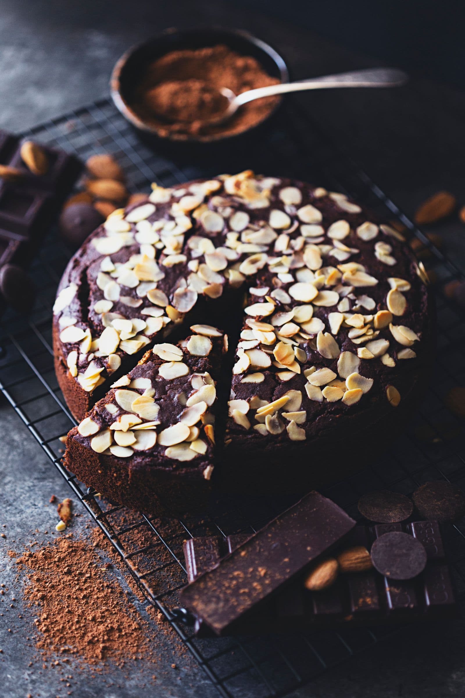 Chocolate almond cake with a slice removed, shown alongside chocolate bars and cocoa powder.