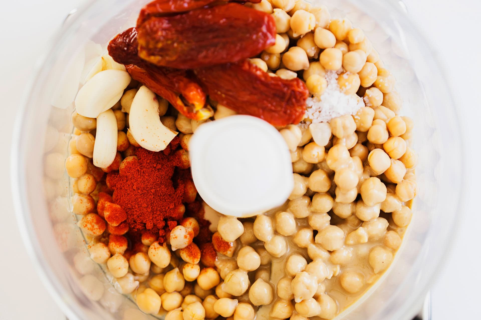 Chickpeas, garlic, tahini, spices, and sundried tomatoes in a food processor bowl.