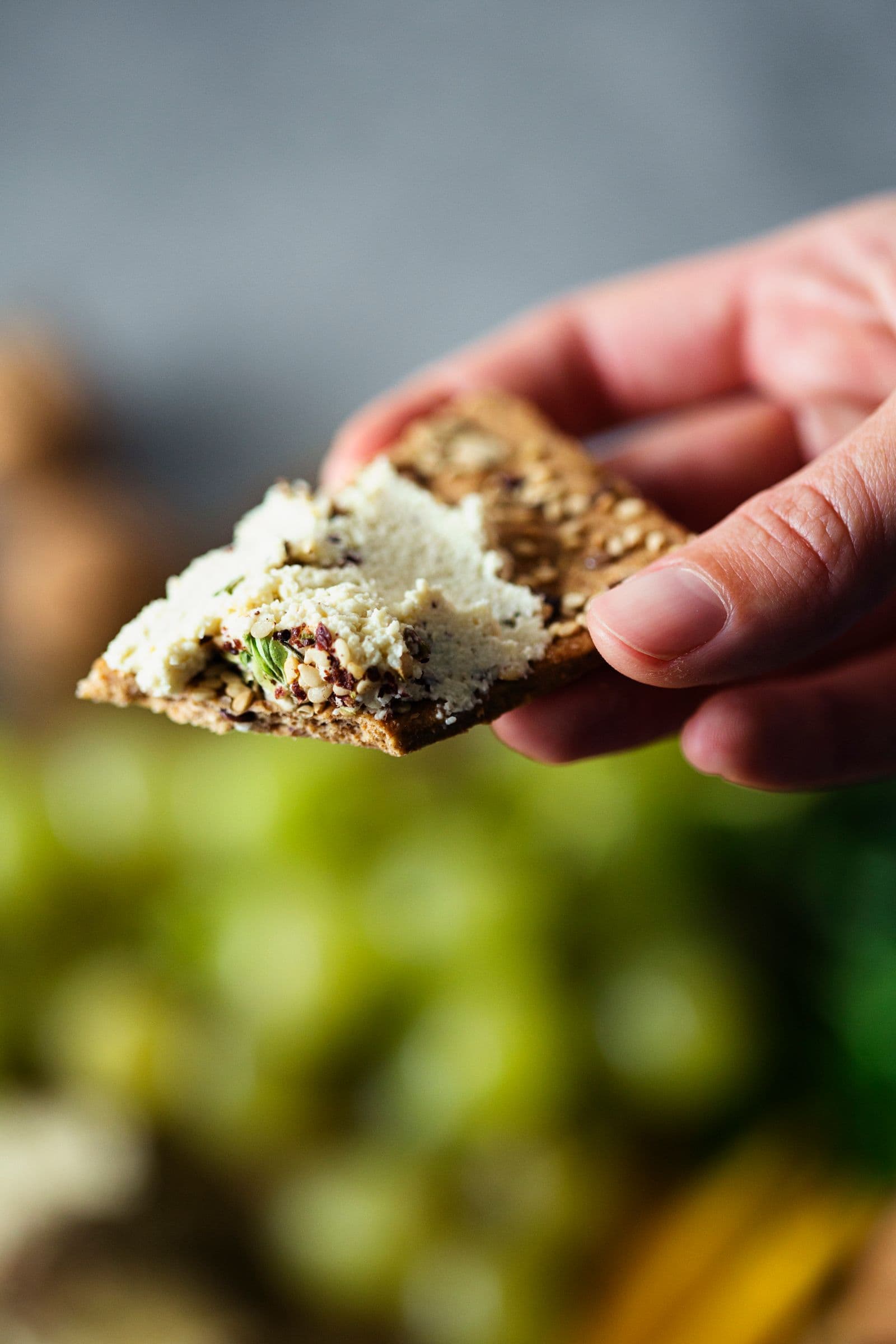 Close-up of hand holding a cracker spread with cashew cheese and fresh herbs.