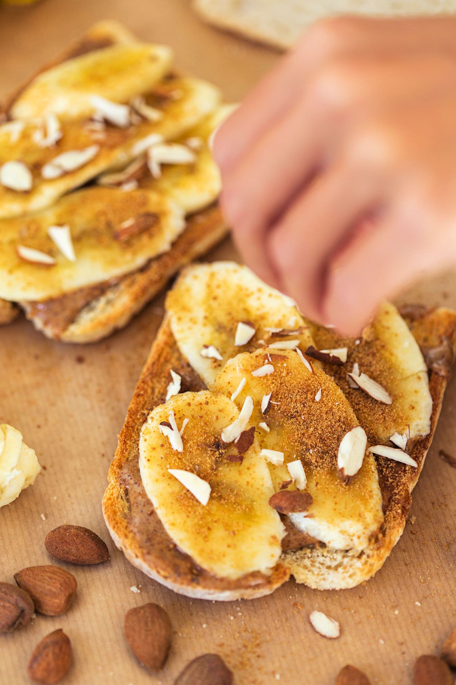 Mão a polvilhar amêndoas picadas sobre uma tosta com manteiga de amêndoa e banana.