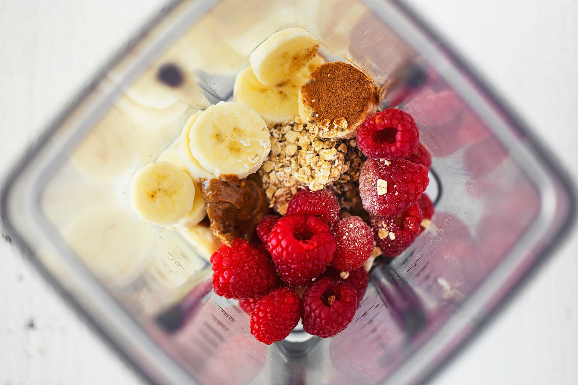 Rodelas de banana, framboesas, flocos de aveia, manteiga de amêndoa e especiarias num copo de liquidificador, prontos a triturar.