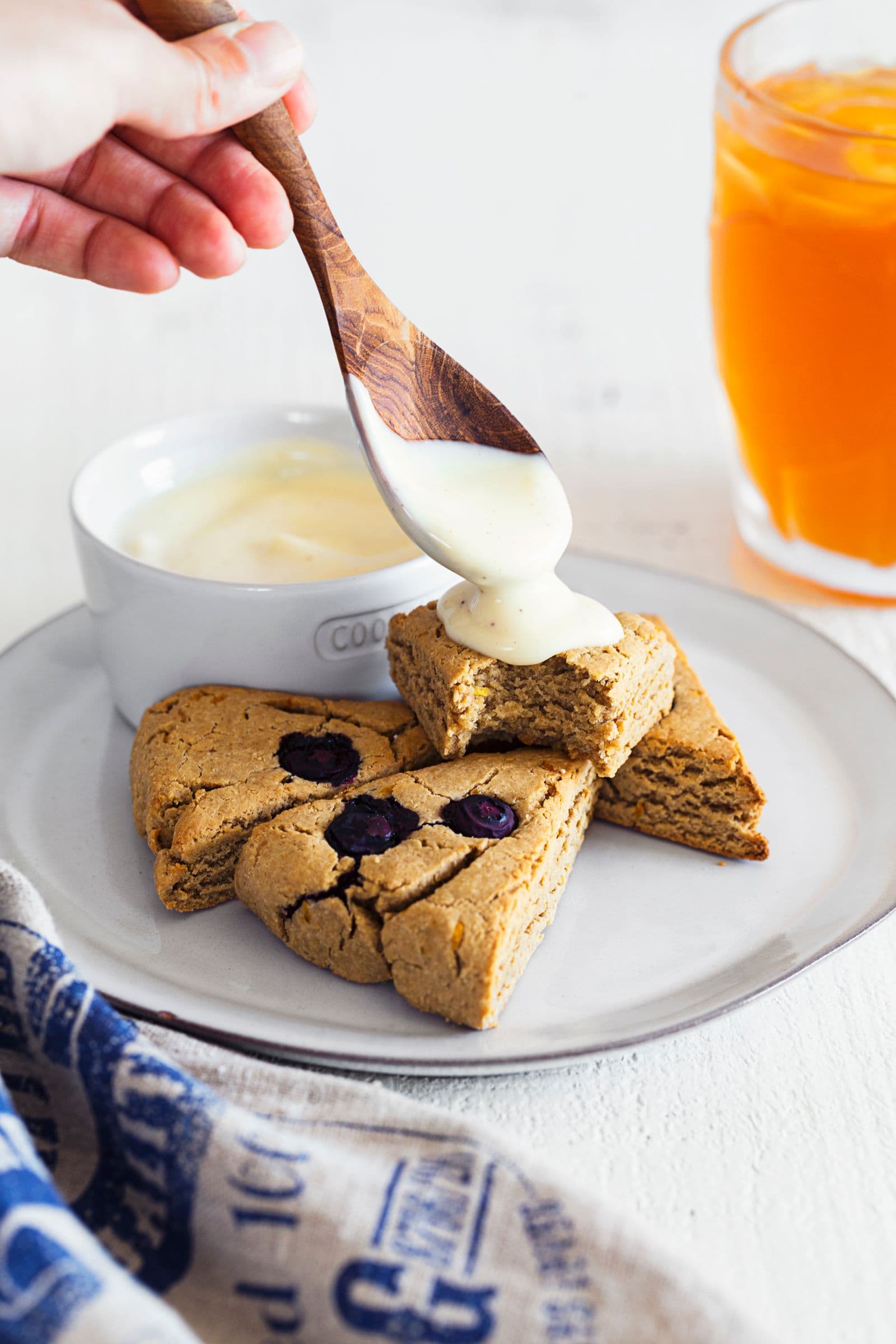 Hand spooning cream over a warm vegan blueberry scone on a white plate.