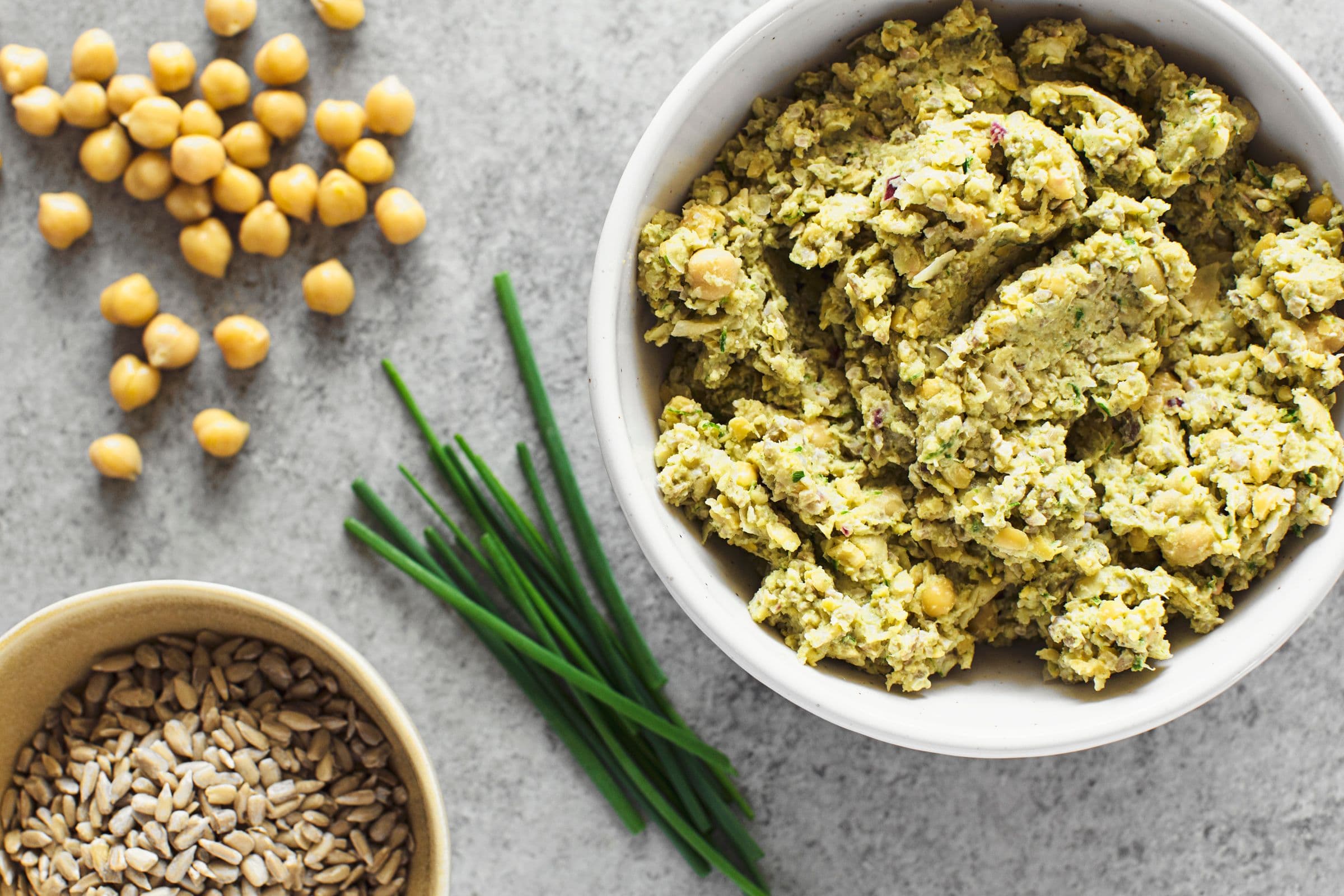Thick chickpea and sunflower seed spread served in a white bowl next to some chickpeas, chives and sunflower seeds..