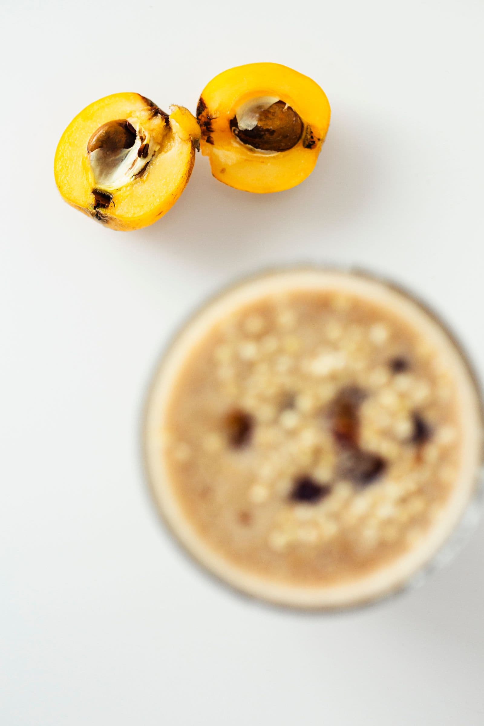 Halved loquat with exposed seeds next to a blurred glass of smoothie topped with puffed millet and a chopped medjoul date