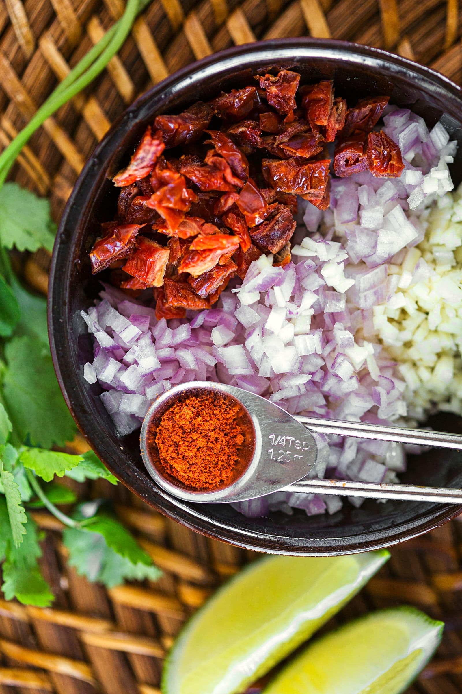 Bowl with chopped sun-dried tomato, onion, lime wedges, and a measuring spoon of paprika.