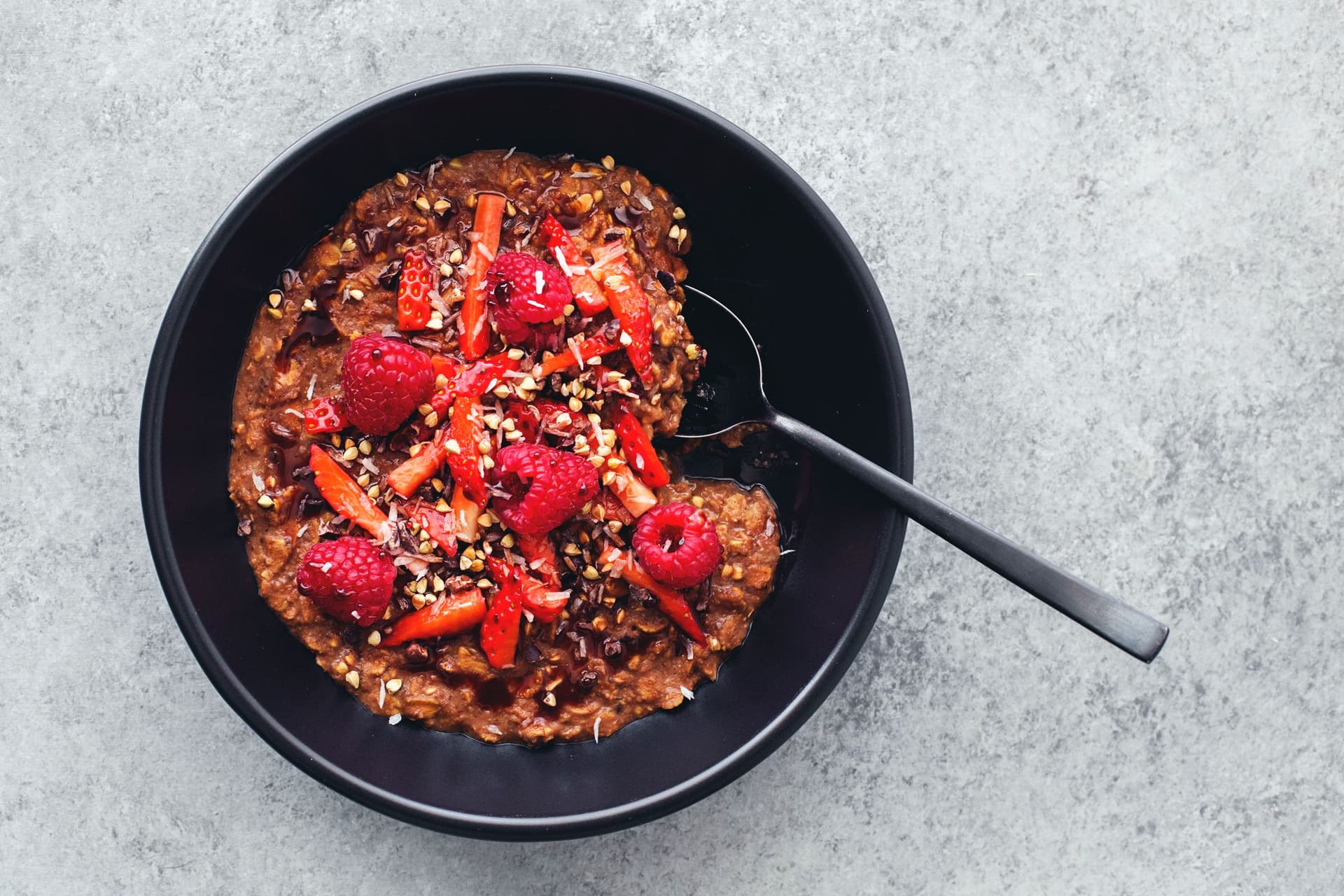 Bowl of cacao oatmeal topped with raspberries, strawberries and crunchy seeds on a grey background.