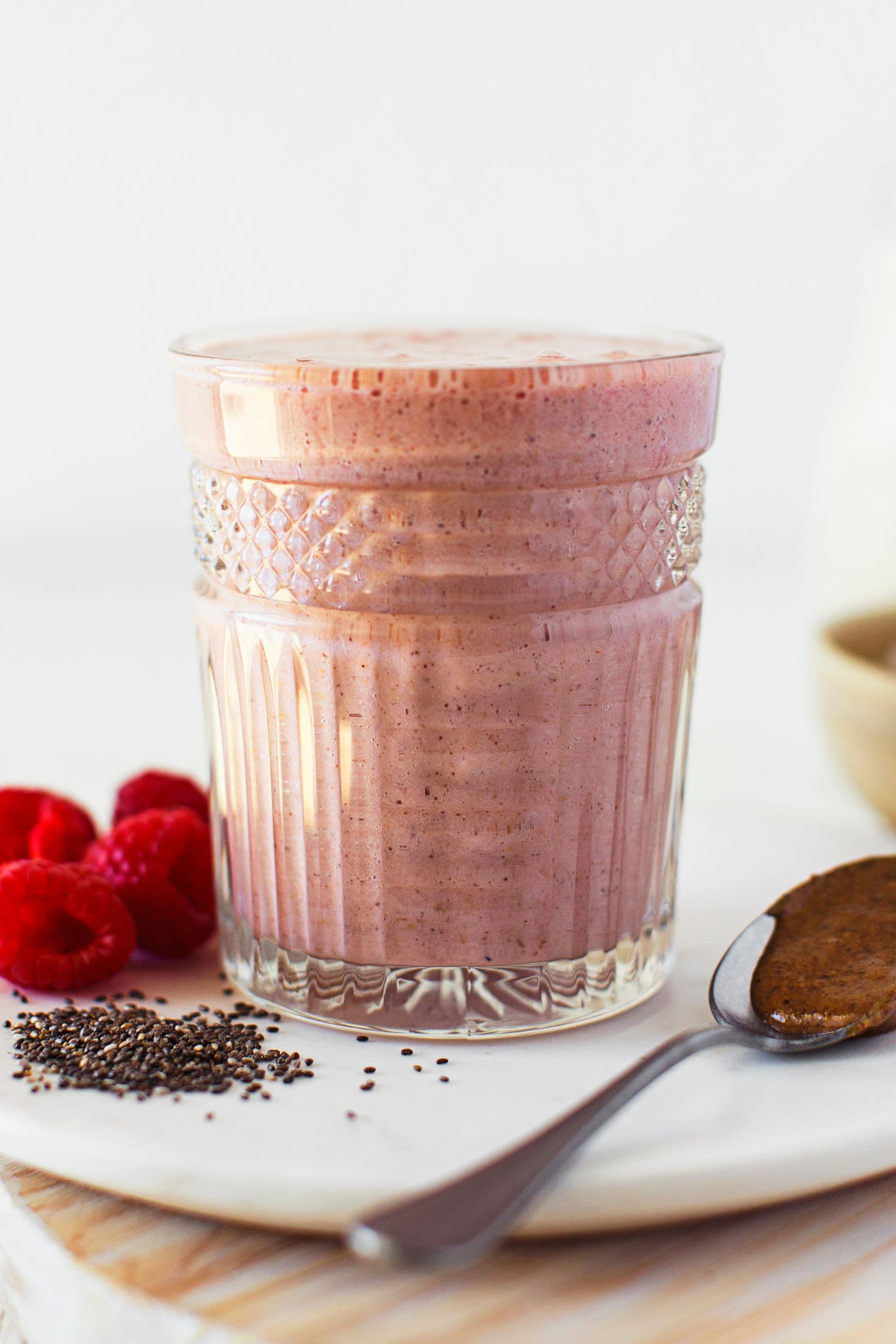Copo com batido rosa sobre um prato, acompanhado de sementes de chia, framboesas e uma colher.
