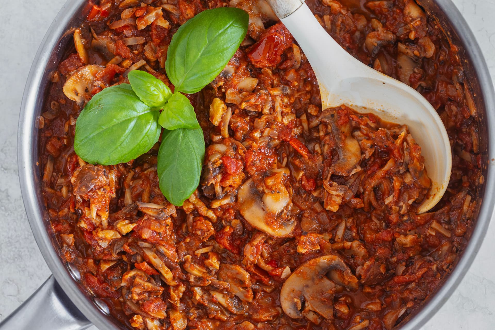 Pot of rich mushroom and walnut bolognese topped with basil leaves and a spoon resting inside.
