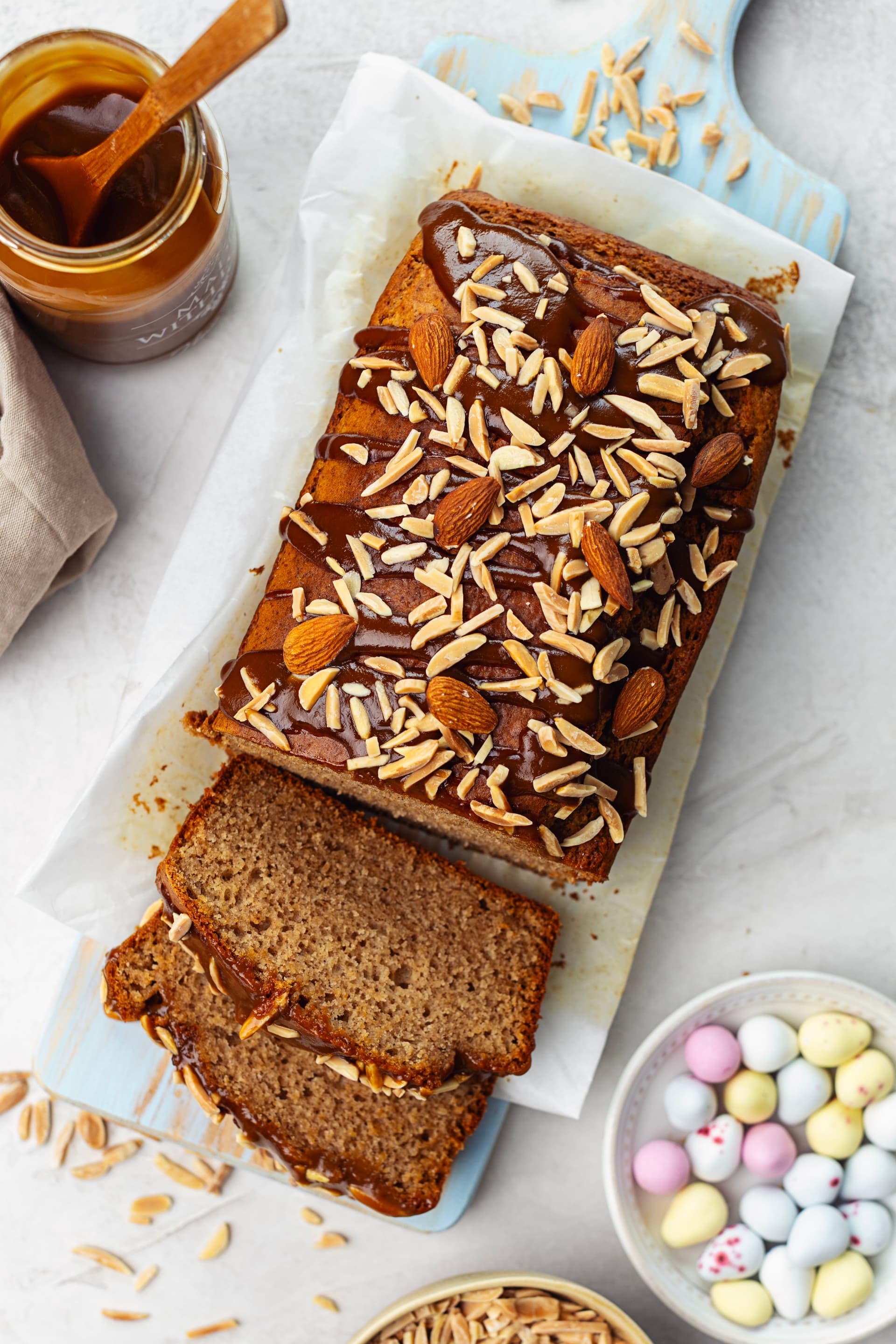 Bolo de amêndoa com caramelo de coco parcialmente fatiado numa tábua azul, com frasco de caramelo e ovos de Páscoa.