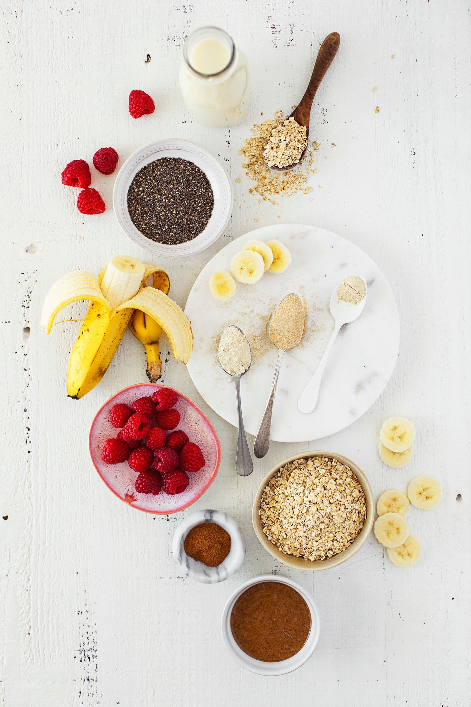 Ingredientes para o batido dispostos numa mesa: banana, framboesas, flocos de aveia, bebida vegetal e superalimentos.