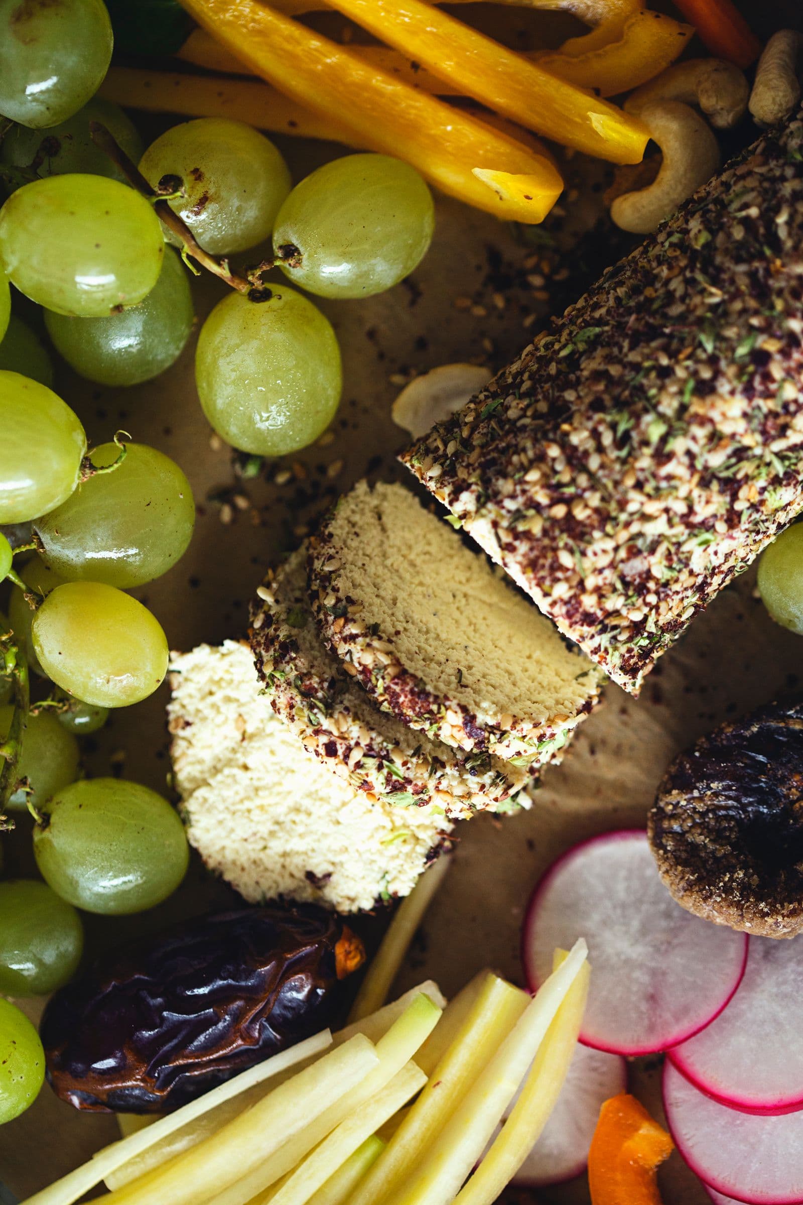 Rolo de queijo de caju fatiado numa travessa, rodeado de uvas verdes e legumes frescos.