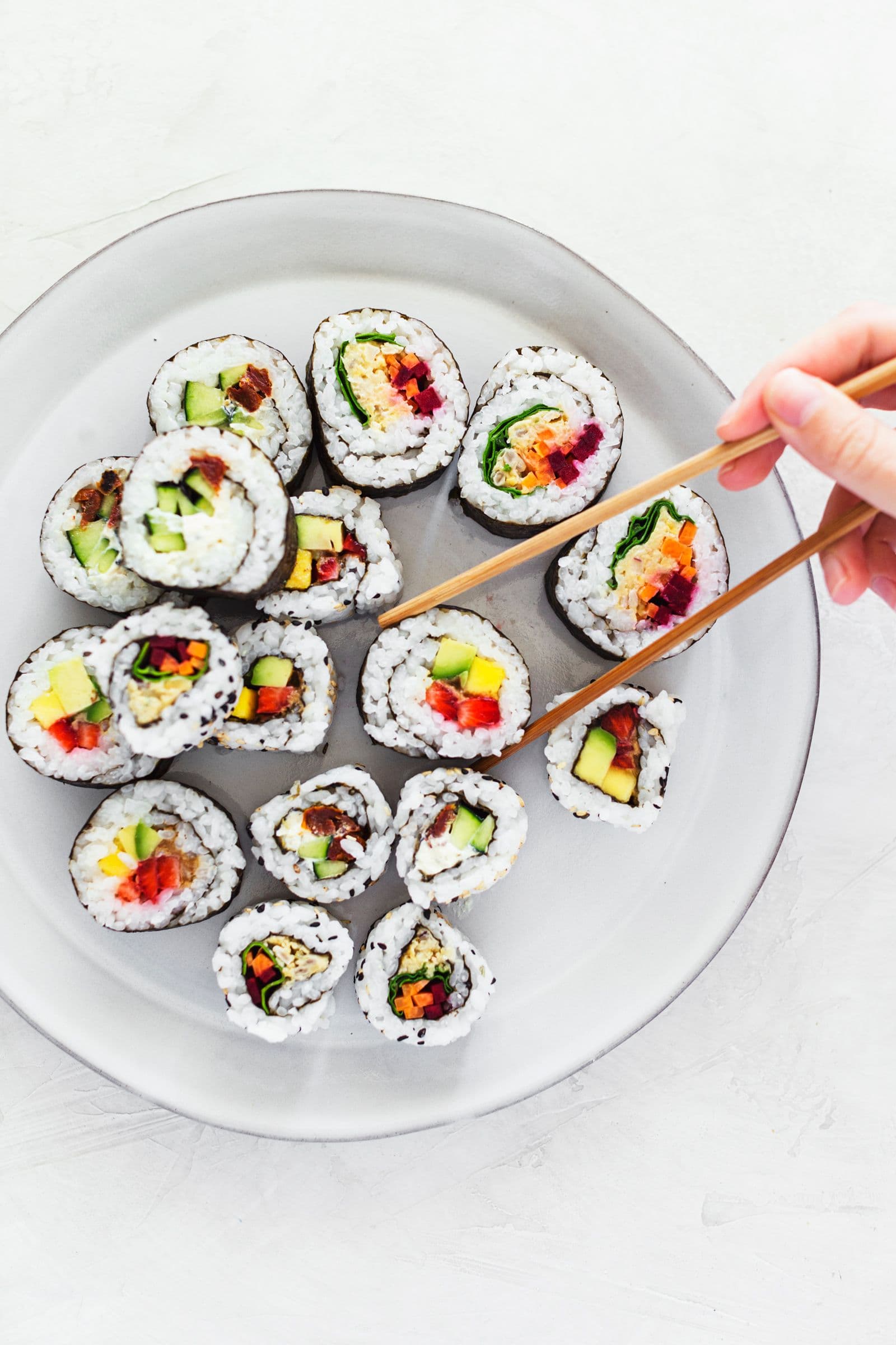 Mão a pegar num pedaço de sushi vegan com pauzinhos de um prato.