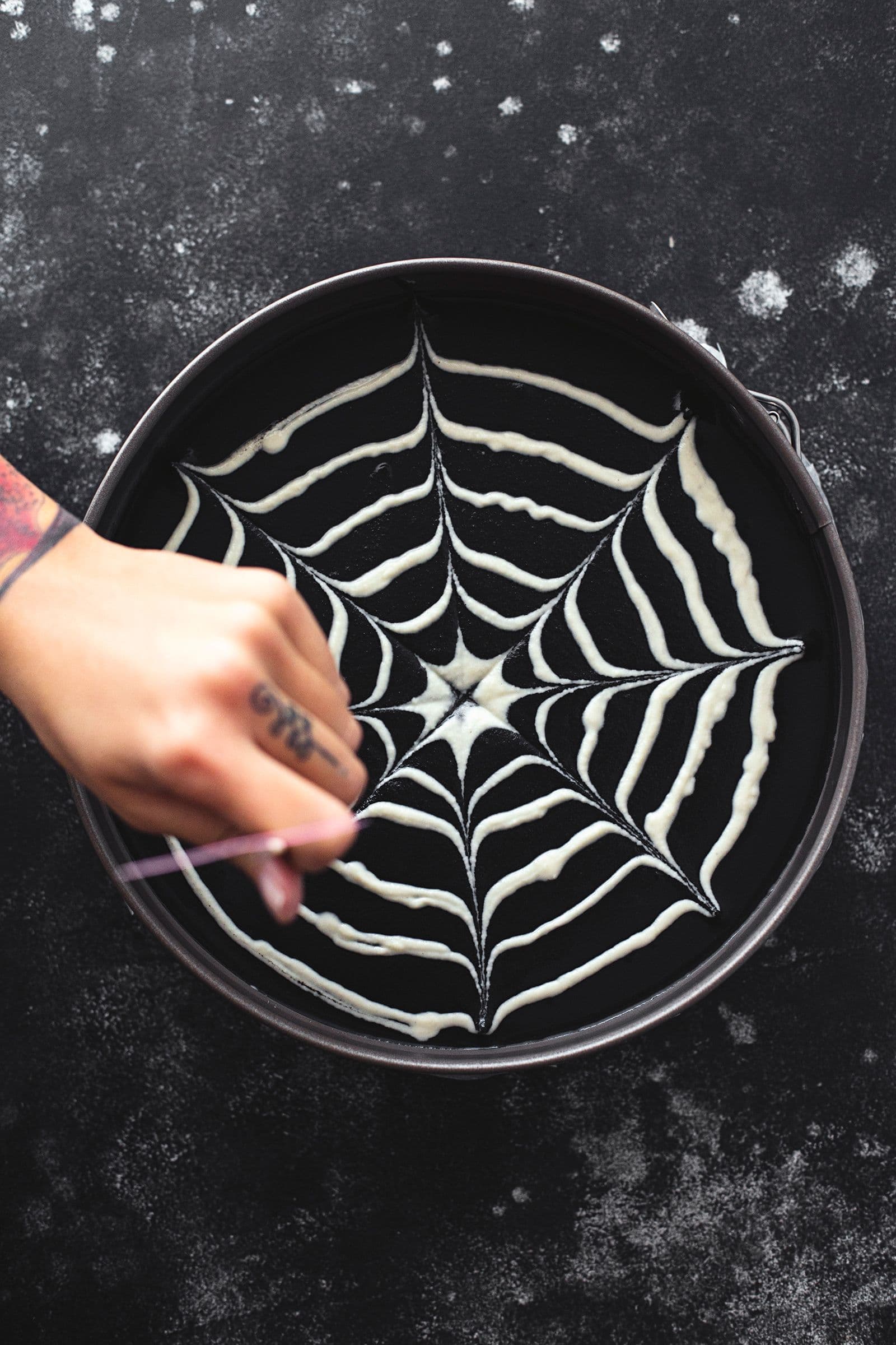 A toothpick dragging through the dots to create a spiderweb design on the cheesecake surface.