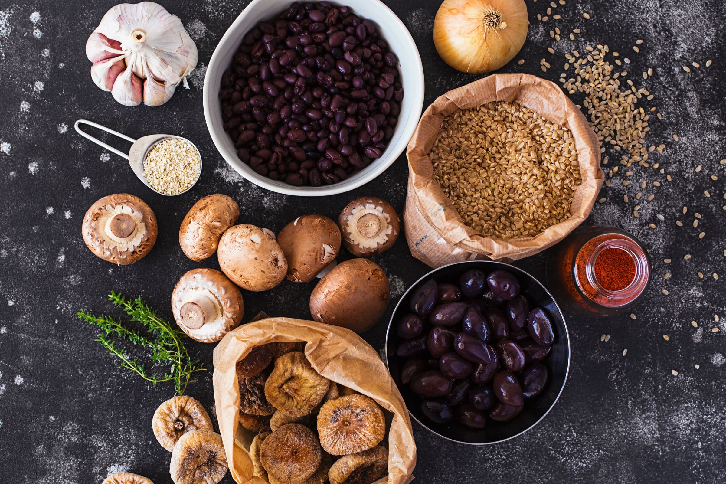 Flat lay of black beans, mushrooms, dried figs, brown rice and olives on a black surface.