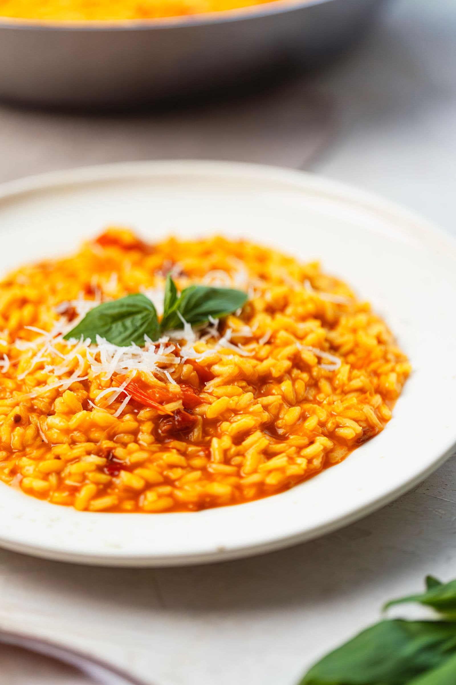 Risotto de tomate cremoso num prato branco, coberto com parmesão ralado e manjericão, com a frigideira ao fundo.