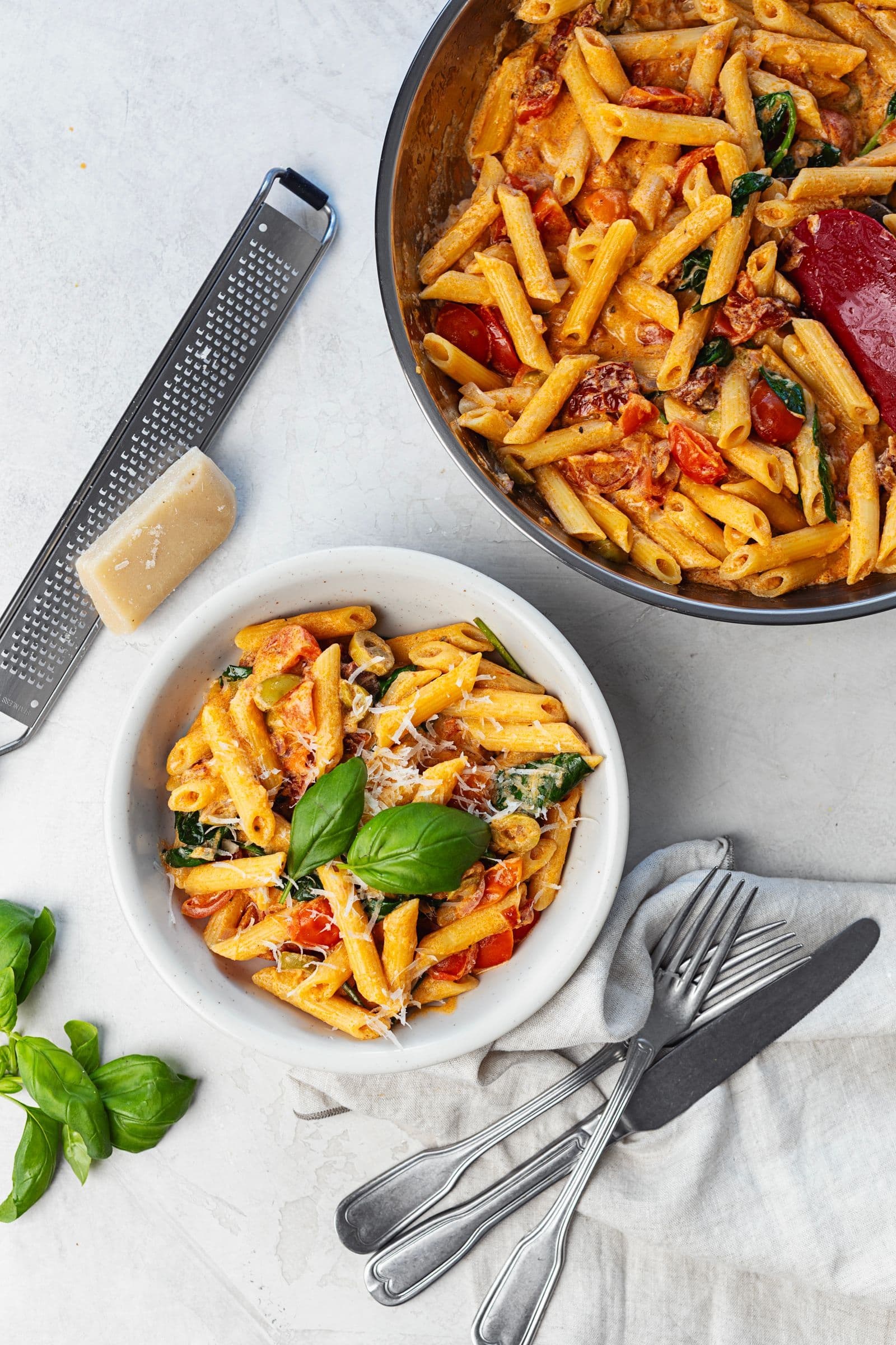 Top-down view of a bowl of creamy tomato and spinach penne next to the serving pan, garnished with fresh basil