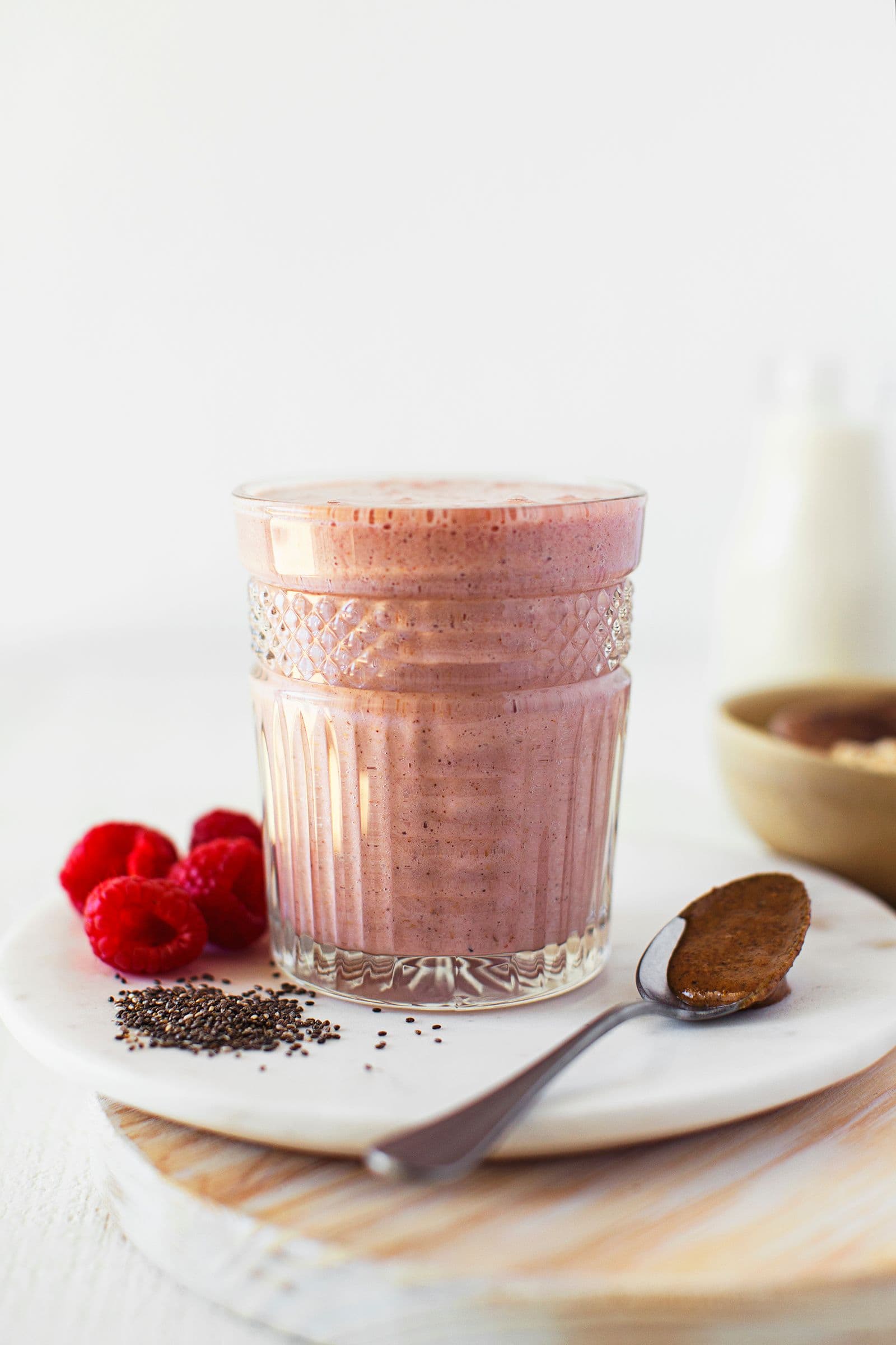 Copo com batido rosa sobre um prato, acompanhado de sementes de chia, framboesas e uma colher.