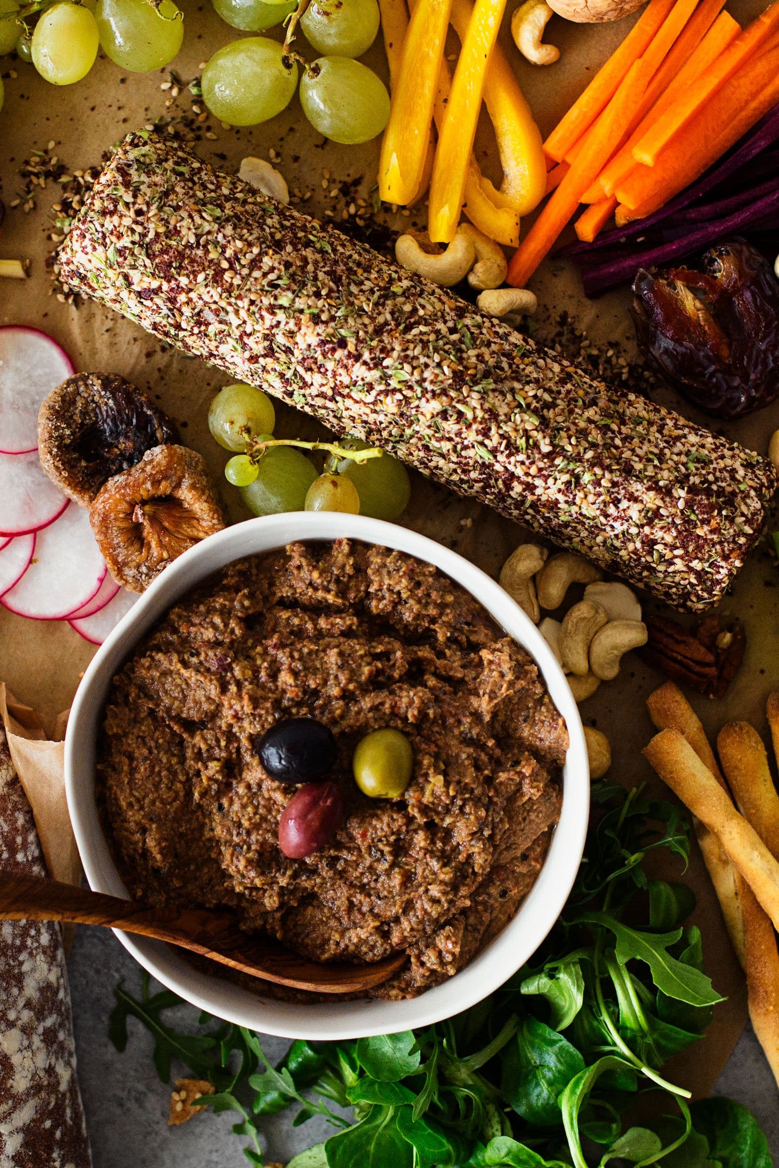 Festive platter with a cashew cheese log coated in herbs, a bowl of lentil and olive pâté, fresh grapes, vegetables and slices of bread.