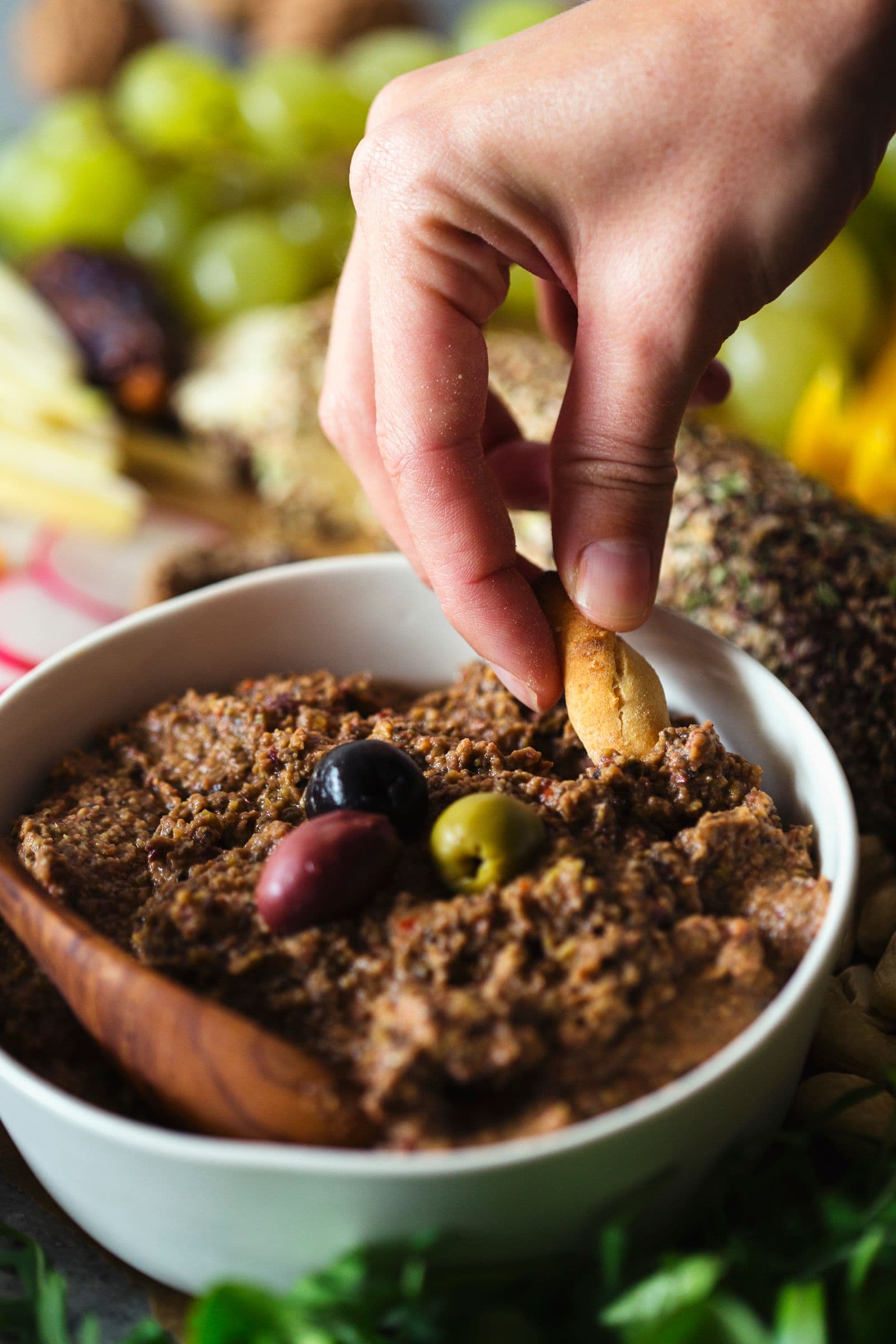 Person dipping bread stick into a bowl of lentil and olive pâté on a sharing platter.