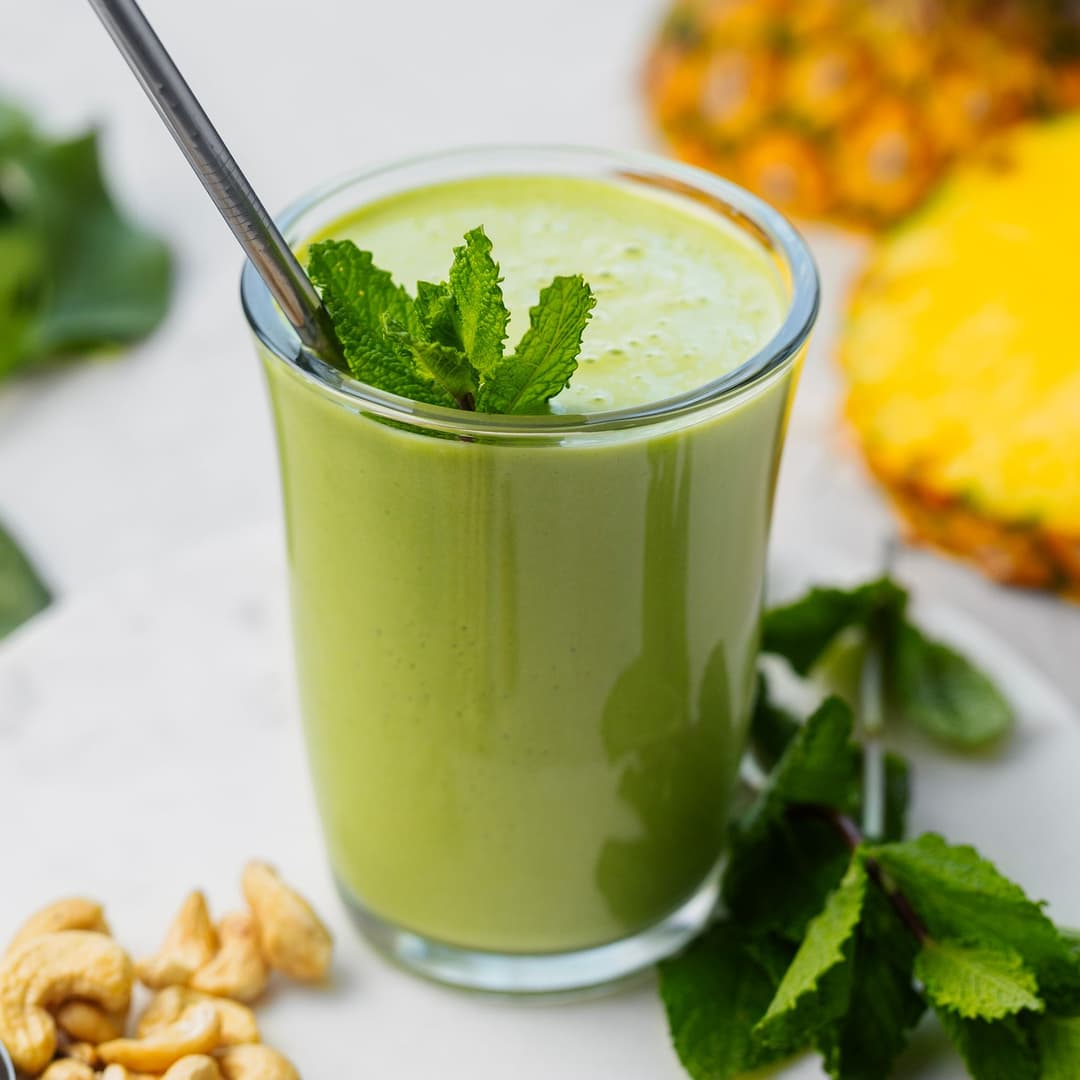 Smoothie verde cremoso num copo com palhinha de metal e hortelã, rodeado por cajus, espinafres e ananás.