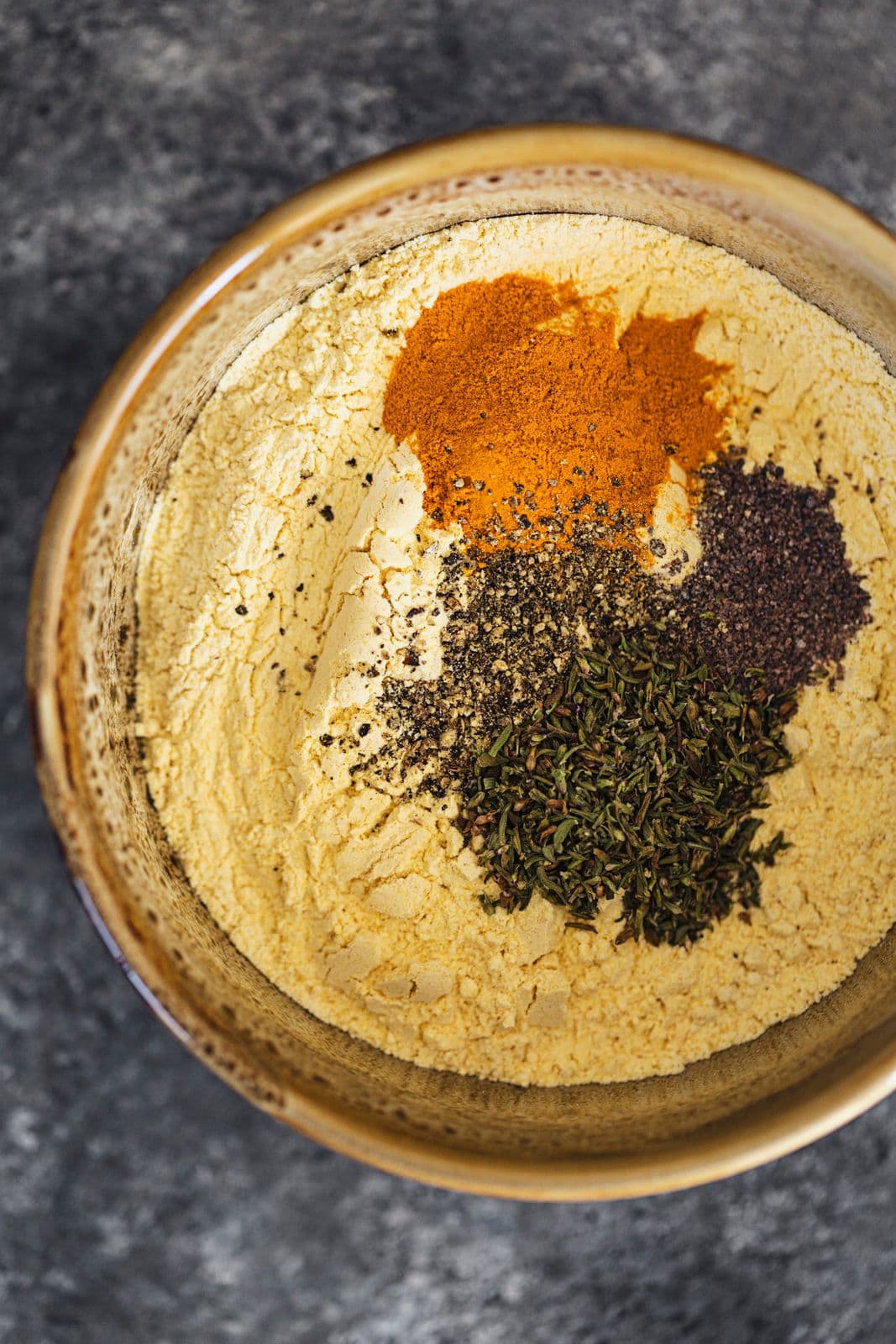 Taça com farinha de grão-de-bico, curcuma, sal, pimenta e ervas secas por cima, pronta a misturar.