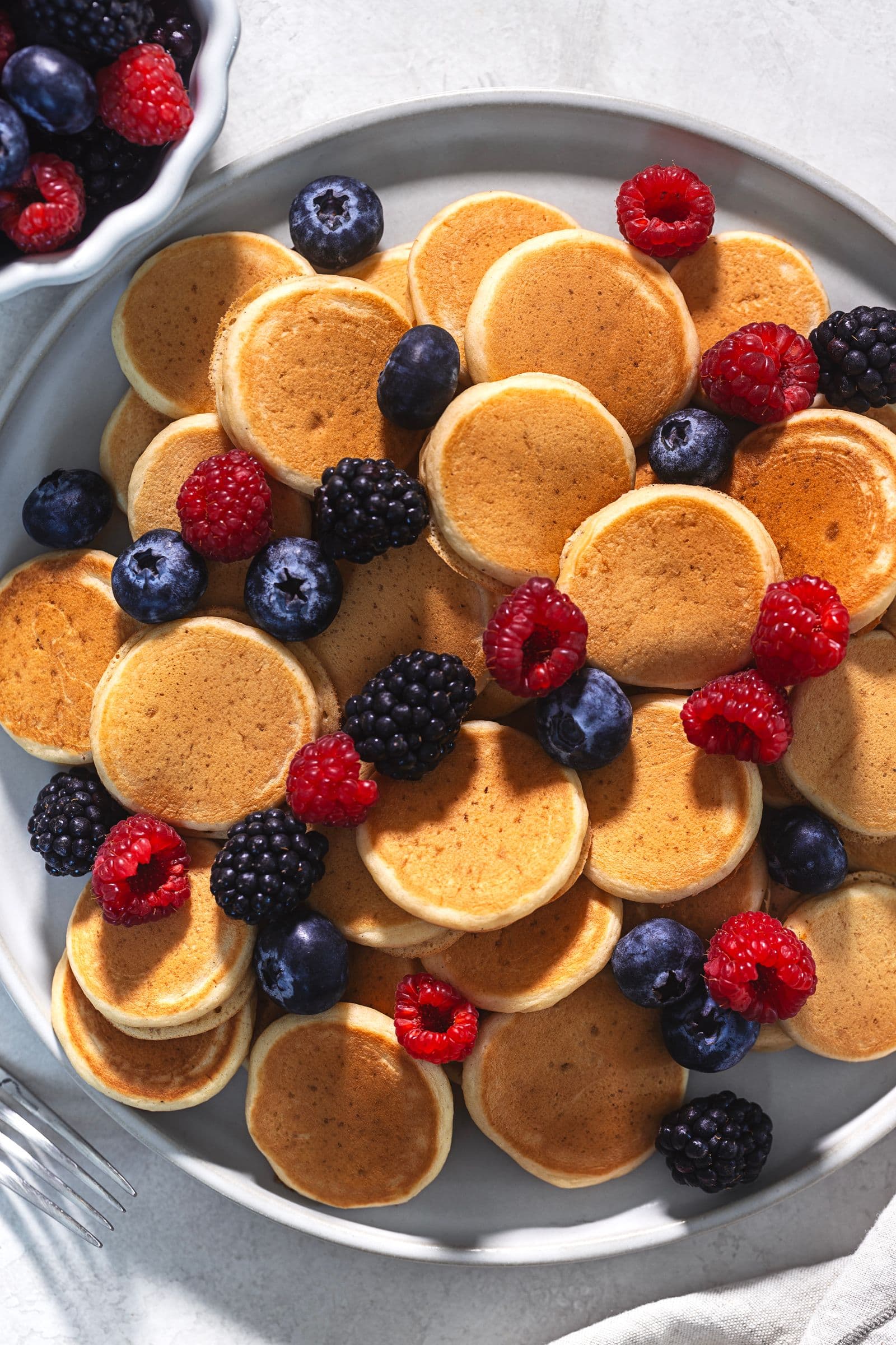 Mini panquecas vegan douradas com frutos vermelhos frescos num prato, com uma taça de frutos ao lado
