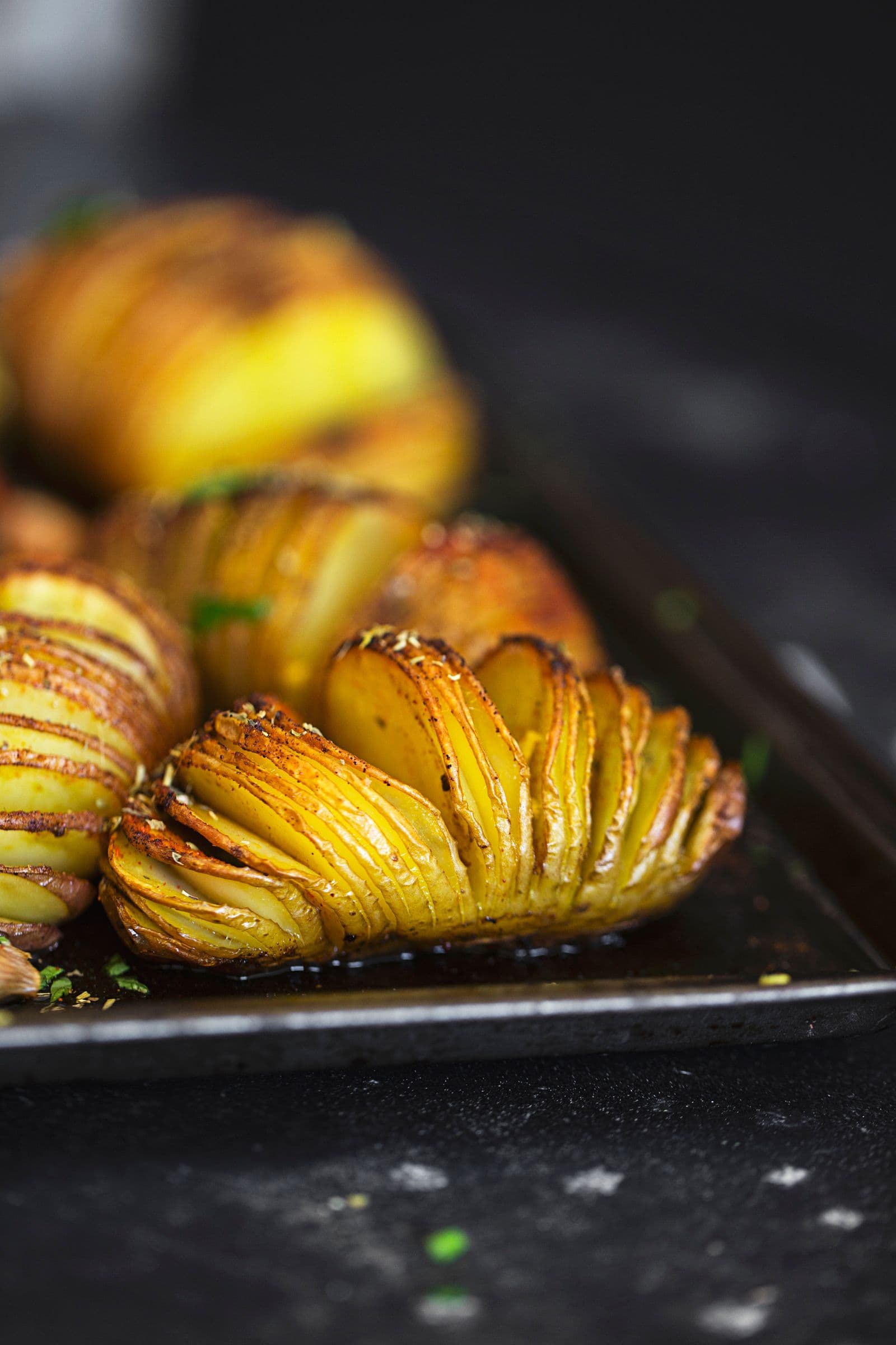 Close view of a seasoned Hasselback potato on a dark baking tray with roasted garlic.