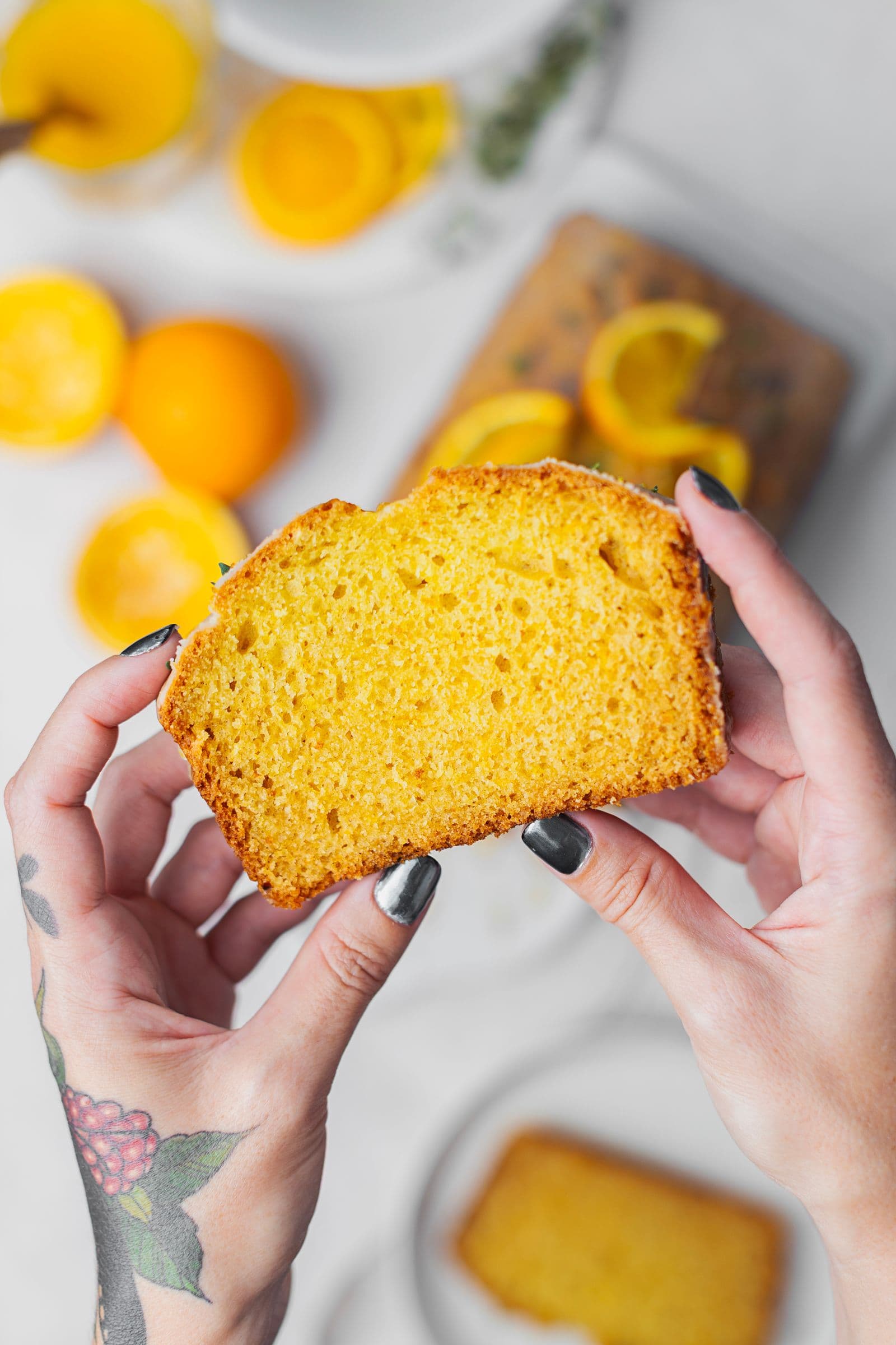 Hands holding a slice of soft orange cake, with blurred background of oranges and the whole loaf.