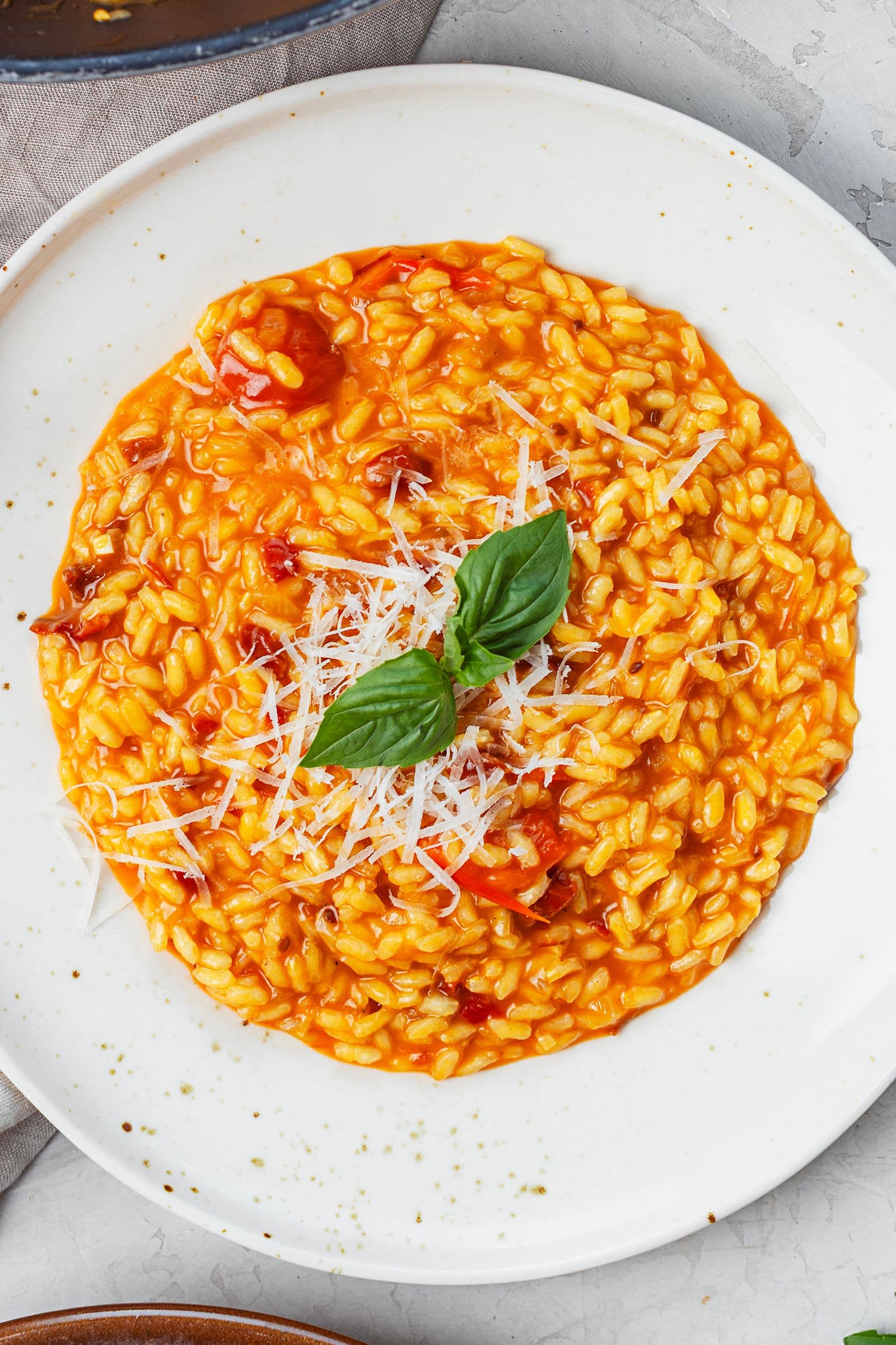 Overhead shot of creamy tomato risotto on a white plate, topped with grated vegan Parmesan and fresh basil leaves.