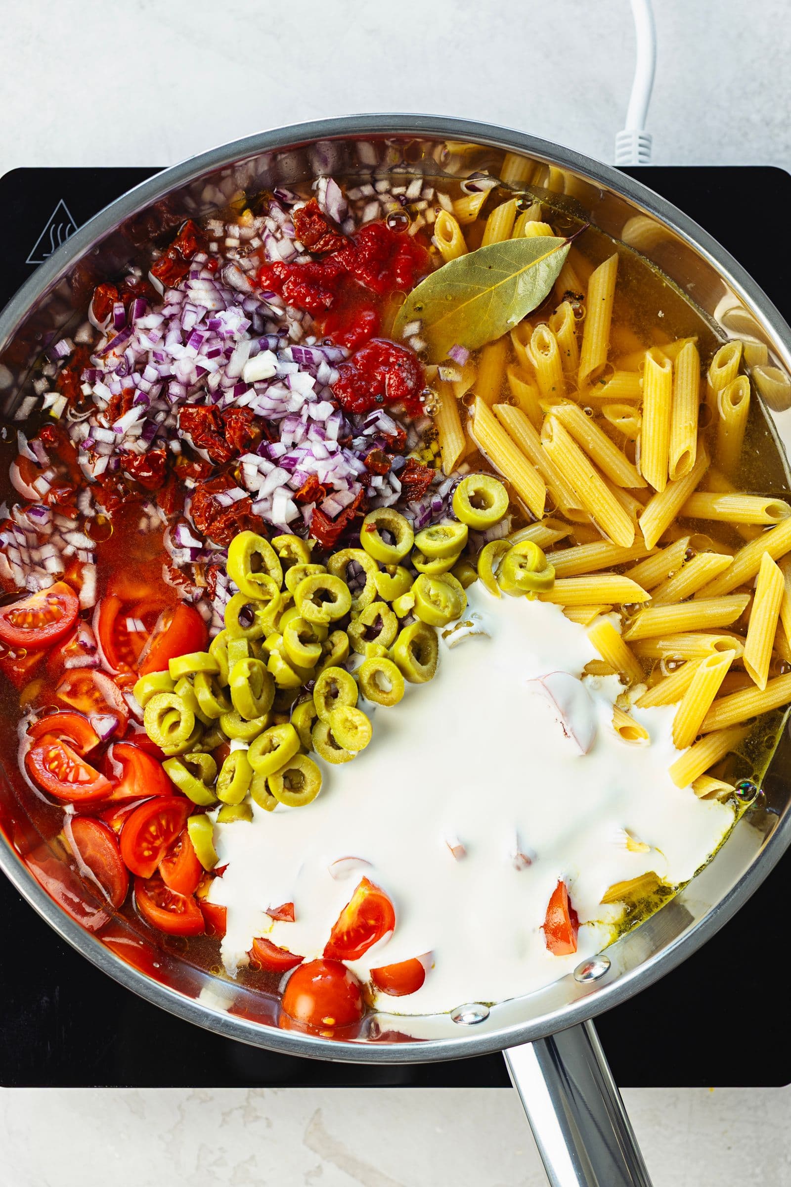 All raw ingredients for one pot pasta added to a large pan, including dry penne, cherry tomatoes, green olives and soy cream