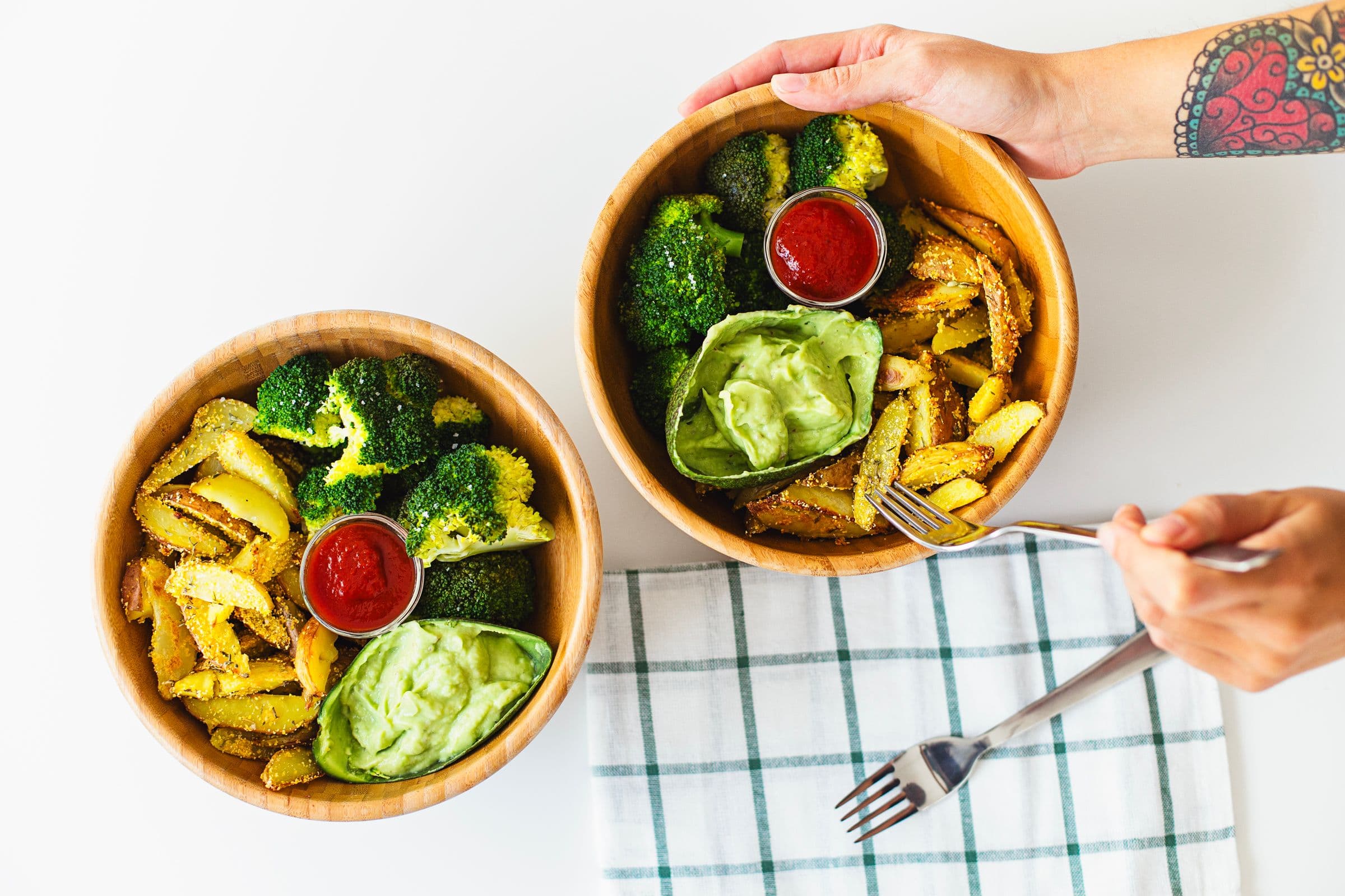 Two wooden bowls with baked wedges, broccoli, guacamole and ketchup being placed on a cloth.
